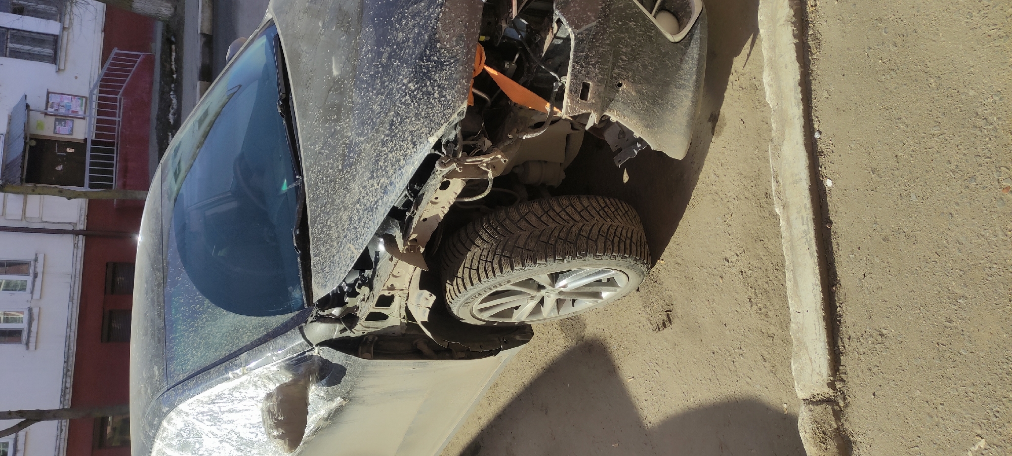 The image depicts a severely damaged dark-colored car, likely in Fryazino, Russia, given the surrounding architecture visible through the windshield. The front of the car is extensively wrecked, with the hood crumpled and missing, exposing the vehicle's internal components. The front left tire is intact and shows a Michelin brand marking. The surrounding environment includes a weathered road and a building with visible windows and a staircase, features typical of urban Russian architecture. The lighting indicates a bright, sunny day. The car's condition suggests a recent collision or impact.