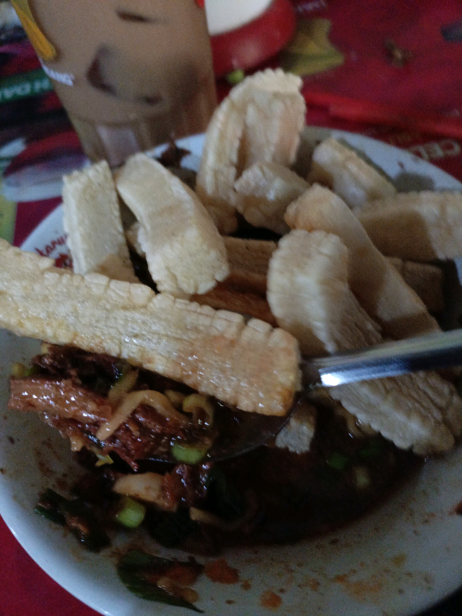 A close-up, overhead shot shows a bowl of Indonesian food in Sraten, Indonesia. The dish consists of a dark, soupy broth with pieces of meat and green onions, topped with several pieces of fried dough. A metal spoon is partially submerged in the broth, scooping up some of the food. The bowl is white and appears to be ceramic. The food is served on a red tablecloth. In the background, a blurred brown beverage in a clear plastic cup is visible. The lighting is dim, suggesting an indoor setting, possibly a restaurant or home. The overall image is slightly blurry and unfocused.