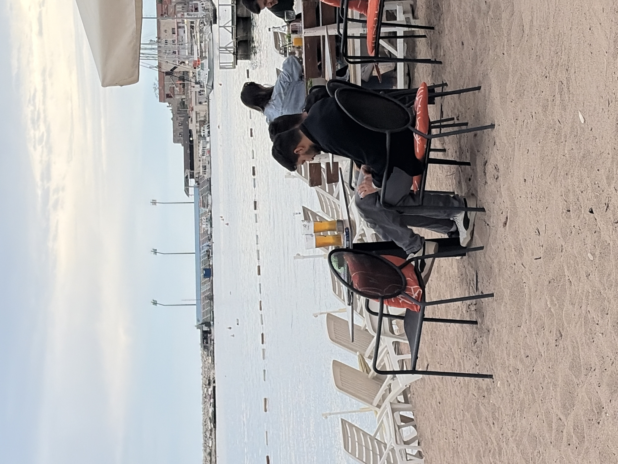 The image depicts a waterfront scene in Budva, Montenegro. Several people are seated at tables under an awning, near a beach. A body of water is visible, with a line of buoys. Beyond the water, buildings and docks are present. Beach chairs are stacked to the side. One man is wearing a black shirt and another person in blue denim. Drinks sit on the tables. The beach is sandy.