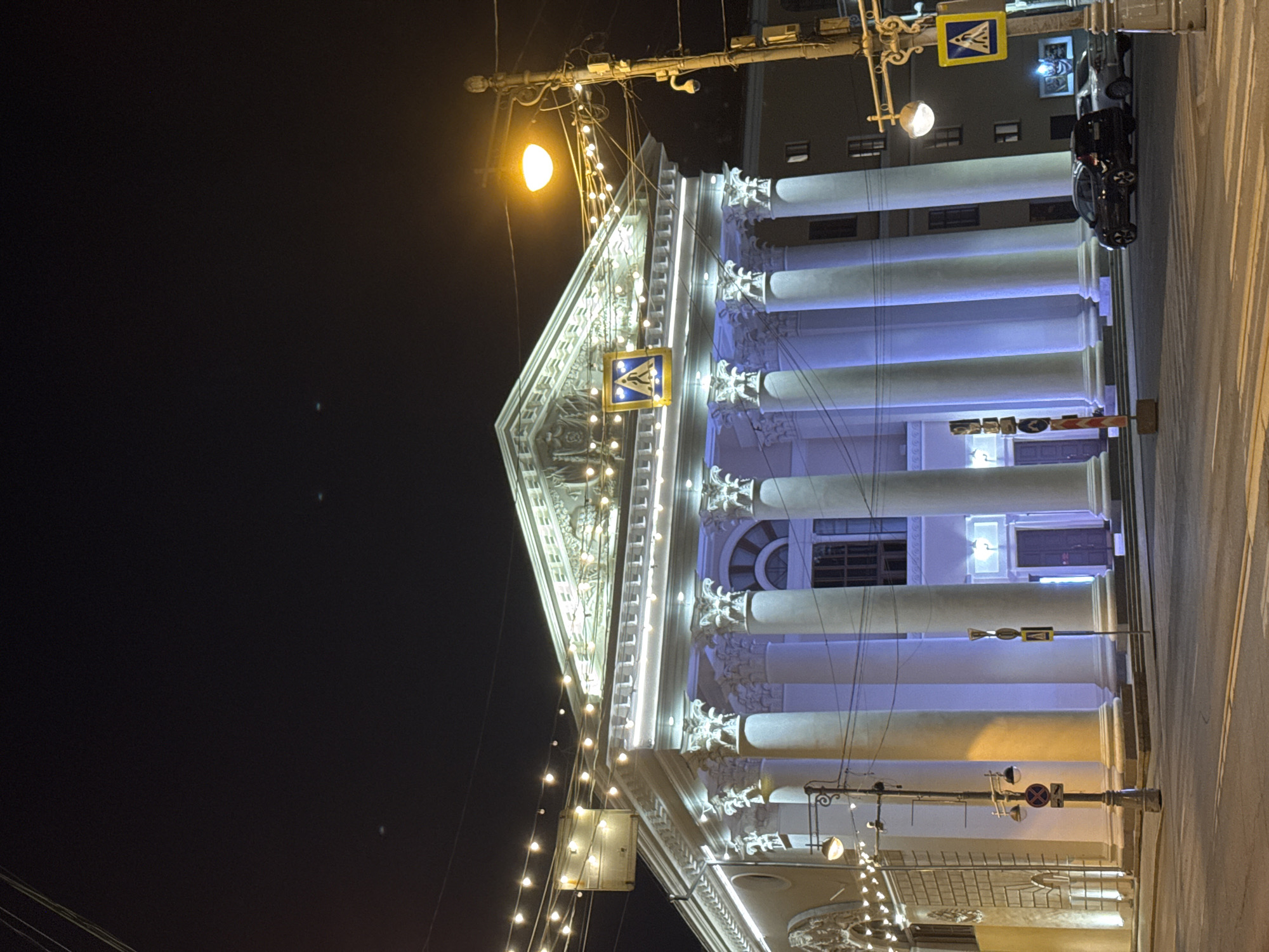 A night-time, low-angle, long shot presents a building illuminated with blue and white lights. The architectural style features a triangular pediment, classical columns, and ornate carvings. A pedestrian crossing sign and a street lamp are visible at the top. A string of lights extends downwards from the apex of the pediment. The building's facade is bright against a dark sky. Cars are parked on the street adjacent to the building. This appears to be the Tver Academic Drama Theater in Tver, Russia.