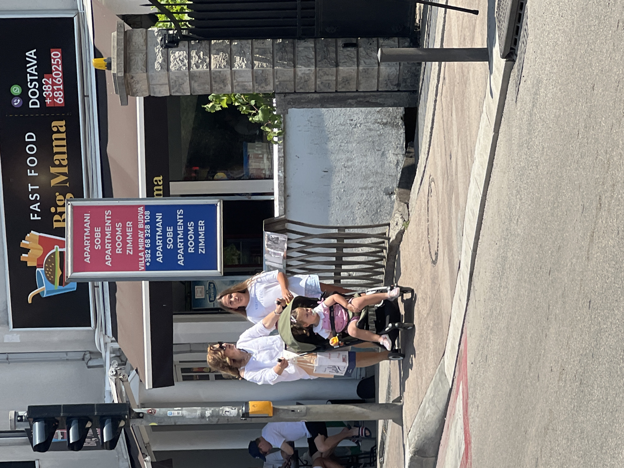 A bright, sunny day in Budva, Montenegro, is captured in this image. A woman in a white shirt and skirt, accompanied by a young girl in a stroller, walks along a sidewalk. The girl is wearing a pink dress and white socks. Beside them, another woman in a white t-shirt and blue shorts walks.
In the background, a "Big Mama" fast food restaurant is visible, with a sign advertising apartments, rooms, and zippers. A traffic light stands on the left side of the frame, and a stone pillar with a black gate is on the right. The environment suggests a busy urban area, likely a commercial street.