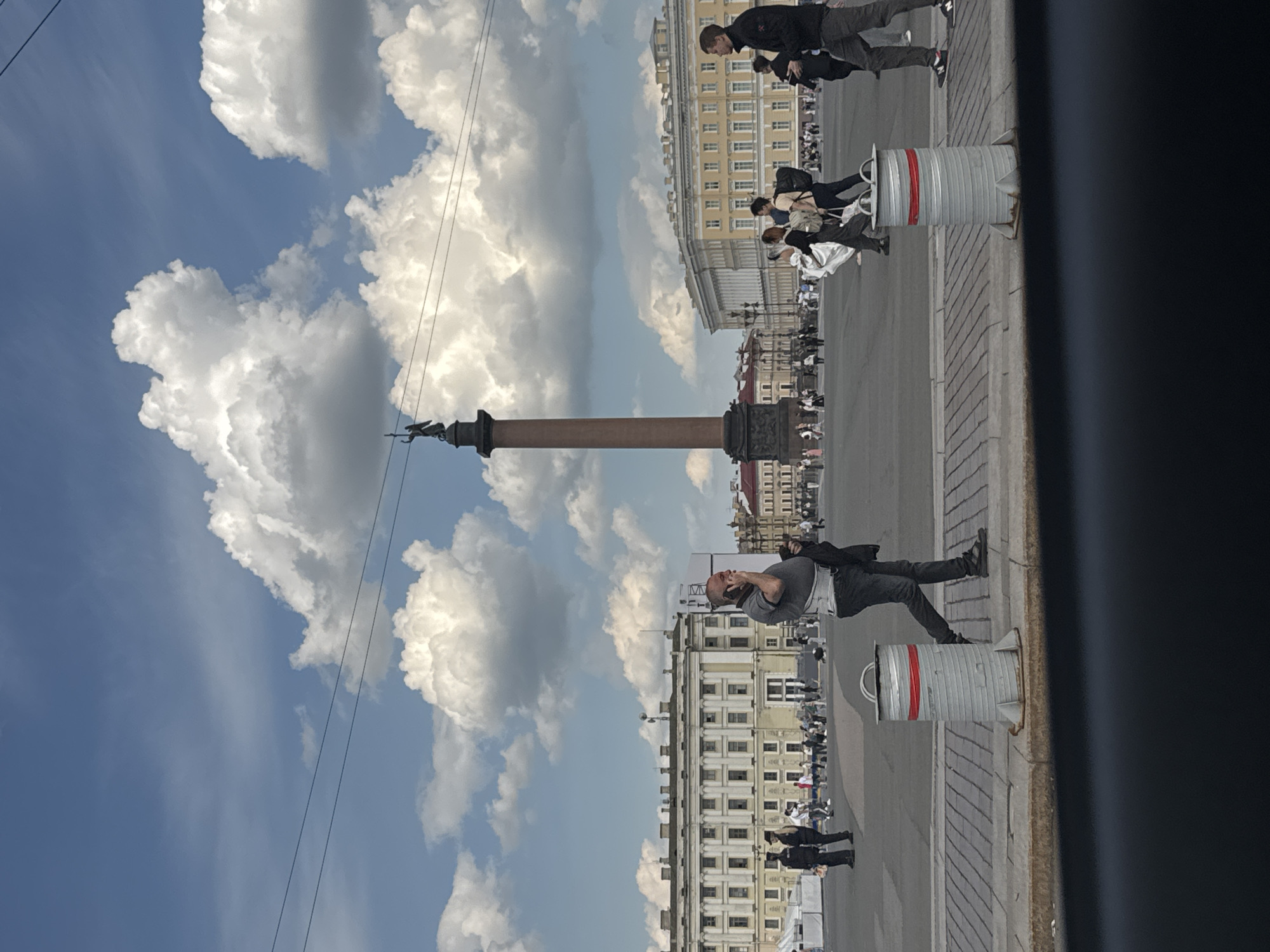 This media file captures a sunny day in a grand public square, likely Palace Square in Saint Petersburg, Russia, centered around the iconic Alexander Column.
The central feature is the tall, reddish-brown Alexander Column, topped with a statue of an angel holding a cross, standing proudly under a bright blue sky dotted with large, fluffy white clouds. Thin power or tram lines are visible crossing the sky.
In the foreground, a man dressed in a grey t-shirt and dark pants stands with his head tilted back, looking upwards, possibly at the column, the statue, or the sky, with his mouth slightly open as if in awe or conversation. To his left, a grey cylindrical bollard with a red stripe marks the edge of the paved area.
Across the square, numerous other people are visible, many strolling or standing. On the left side of the frame, a group of individuals in casual clothing, including a man in a dark jacket and jeans, and another person in a white top, can be seen. Further back, more people mill about, some near the historical buildings that line the square. These multi-story structures feature intricate facades and numerous windows, with a prominent yellow building visible behind the column.
The ground consists of patterned stone paving (sidewalk) and a dark asphalt road. The bright illumination and distinct shadows indicate it is daytime, likely midday or early afternoon, with clear and pleasant weather. No discernible text is visible in the image. The overall atmosphere suggests a typical day of sightseeing and public activity in a historic urban setting.