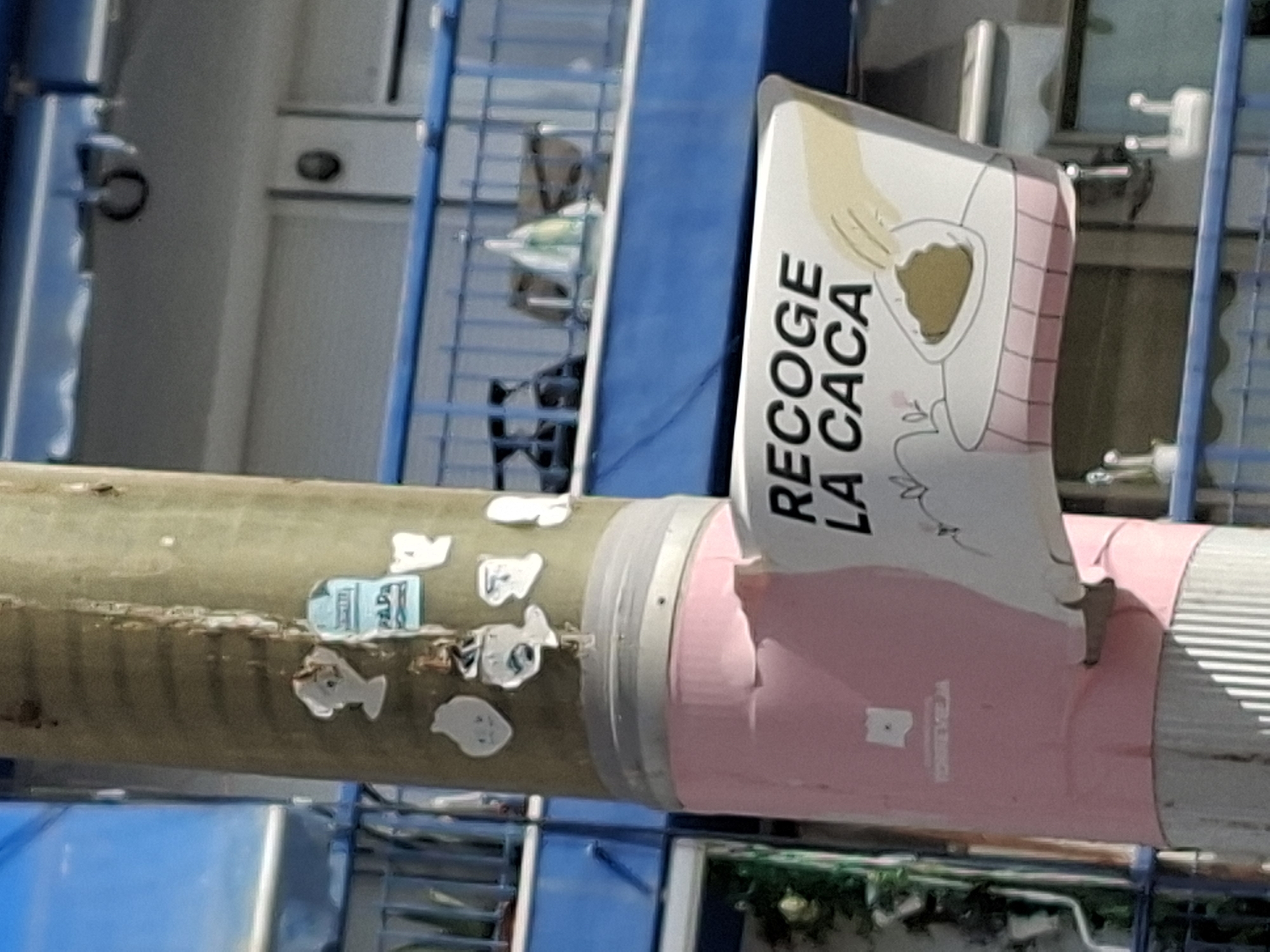 A utility pole in Torrevieja, Spain, is covered in peeling pink tape and various stickers. A sign is affixed to the pole that reads "RECOGE LA CACA," which translates to "PICK UP THE POOP." The sign features a cartoon illustration of a hand scooping dog feces into a bag. In the background, a blue balcony railing and part of a building are visible.
