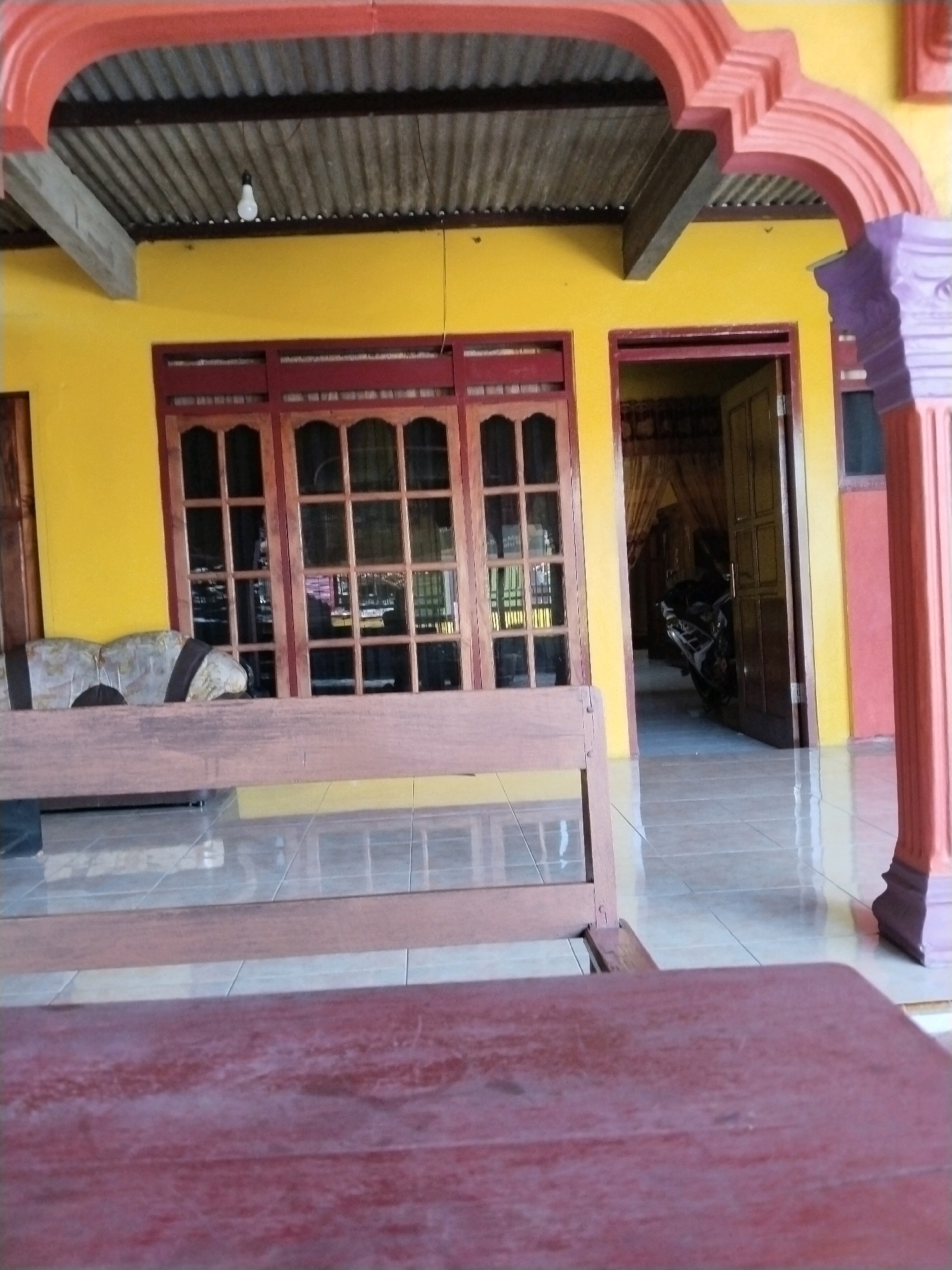 The image depicts the exterior of a building with a yellow facade, accented by a red-orange archway and a purple column. A corrugated metal roof is visible above the yellow wall. In the foreground, a wooden bench is present, with a darker wooden surface in front of it, possibly another bench or table. To the left of the bench, a wooden window with multiple panes is visible, with what appears to be a stone sculpture peeking over the back of the bench. To the right, an open doorway reveals a motorcycle parked inside a dimly lit room with yellow walls and curtains. The floor is tiled with a reflective surface. The scene suggests a residential or commercial property in a warm climate. No people are visible in the image.