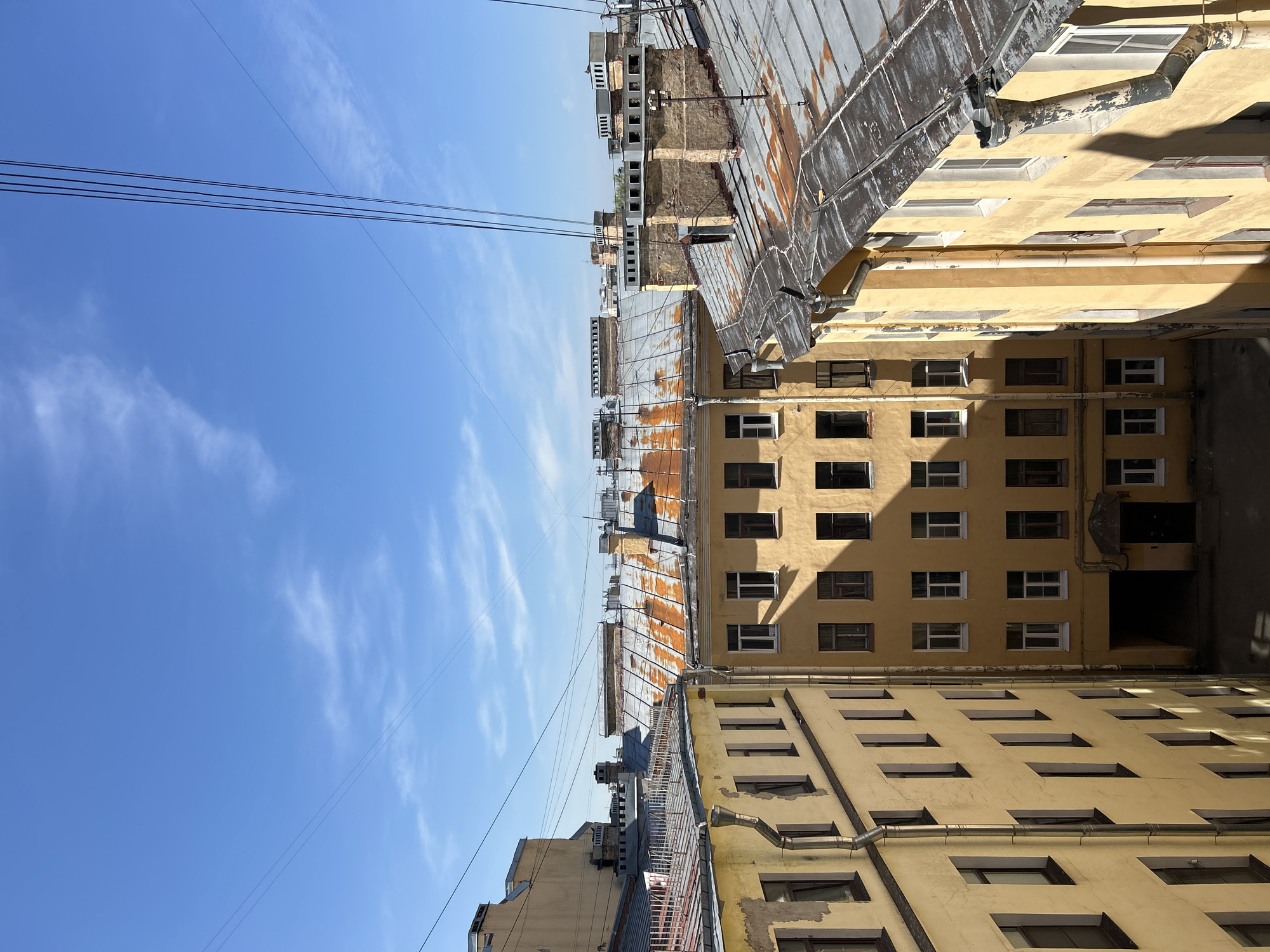 A vertically oriented, eye-level shot captures a narrow courtyard between buildings in Saint Petersburg, Russia. The sky above is a clear blue with wisps of cirrus clouds. Four parallel power lines stretch across the upper-left quadrant of the frame. The buildings are densely packed, with the closest edifices sporting light yellow facades punctuated by rows of rectangular windows. The roofs are tin, corroded with rust. The courtyard is filled with light.