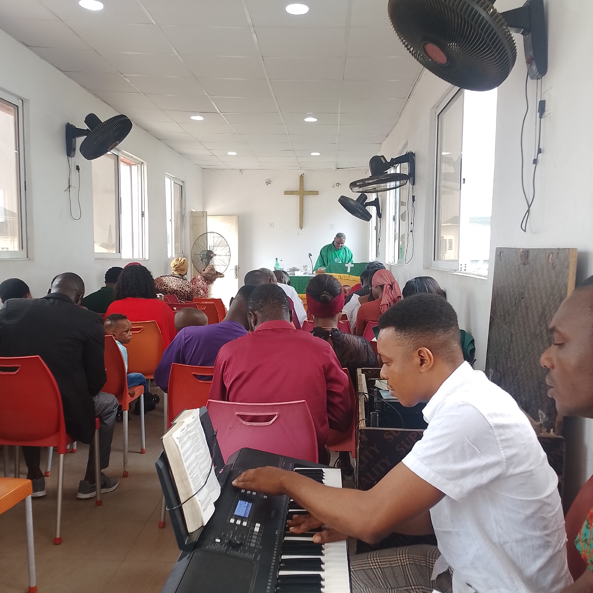 This image captures an indoor gathering, possibly a church service, in Iranla, Nigeria. The room has white walls and a tiled ceiling with several recessed lights. Two large black wall-mounted fans are visible, one on the left side of the room and another on the right. In the background, a wooden cross is mounted on the wall above a podium draped with a green cloth. A person wearing a green robe is standing behind the podium, seemingly addressing the attendees.
The seating area is filled with people, mostly facing towards the front of the room. The chairs are bright orange. In the foreground, a young man in a white shirt is playing a black keyboard. An open Bible rests on a stand next to the keyboard. Another person, partially visible on the right, is looking towards the keyboard player. The floor appears to be tiled with a light brown color. Various individuals in the congregation are dressed in a mix of casual and formal attire, with some women wearing headscarves. The overall atmosphere seems to be one of a community event or religious service.