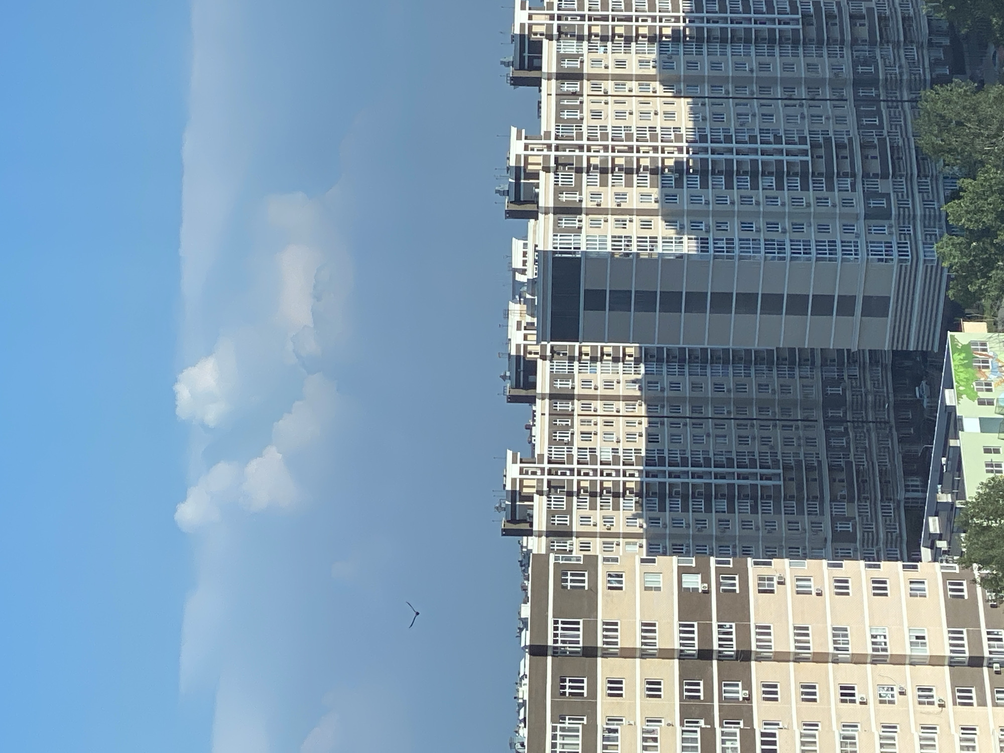 A wide shot captures a modern city skyline under a bright blue sky with scattered white clouds. Multiple tall, multi-story residential buildings dominate the lower half of the frame. These buildings are characterized by their geometric designs, featuring numerous windows and balconies in shades of beige, brown, and white. One particularly striking building in the foreground has a vertical, grey and beige striped pattern. A single bird is visible in flight against the blue sky, high above the buildings. The cityscape is dense, suggesting an urban environment. The overall impression is one of a sunny day in a developed urban area.