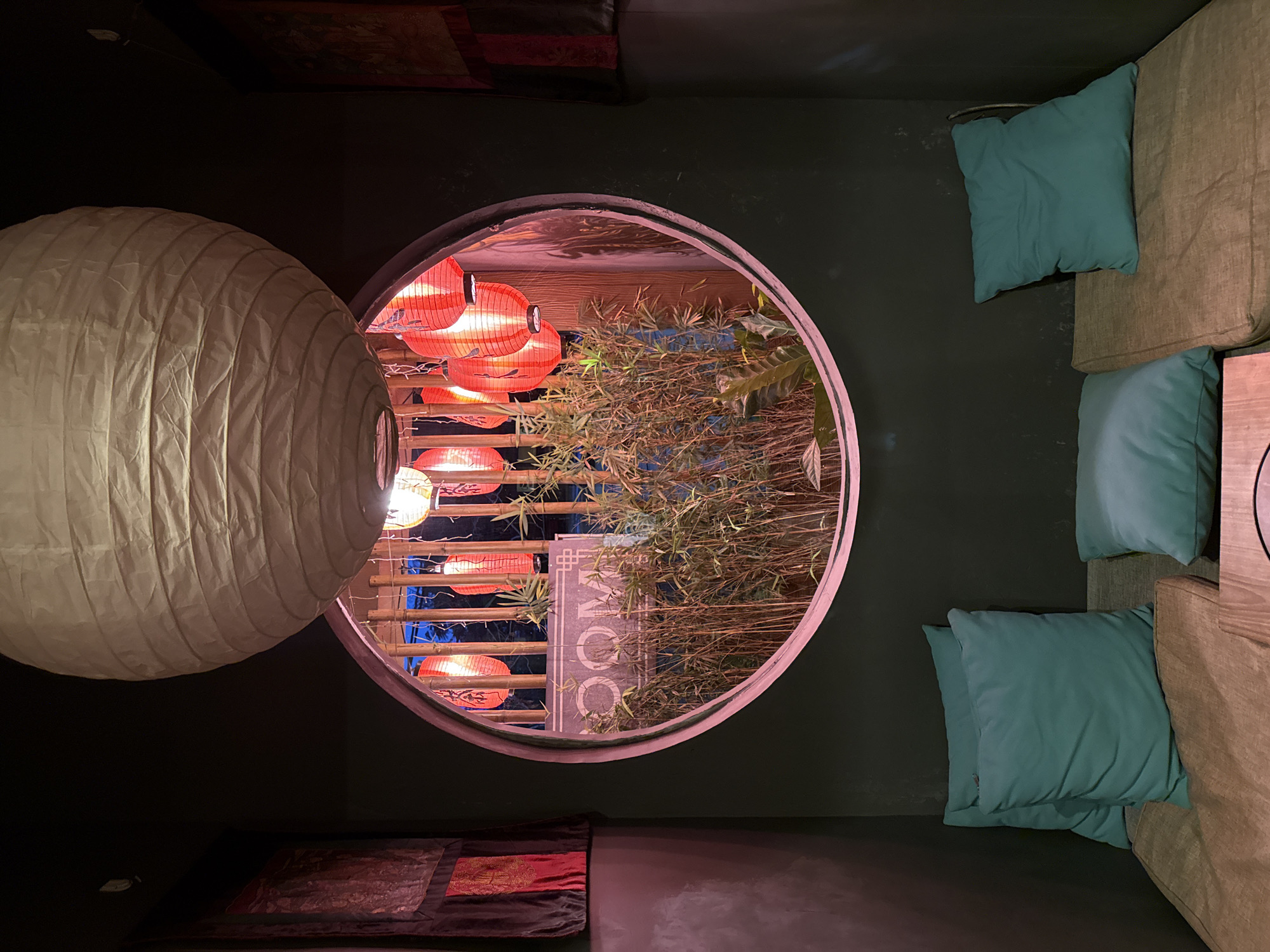 The interior of a building in Kerobokan Kelod, Indonesia, is depicted. A large, round window frames a scene with red lanterns and bamboo. A paper lantern hangs on the left, partially obstructing the view through the window. To the right of the window are cushions and a sofa. Above and below the window are tapestries or cloths hanging on the wall. A sign can be seen inside the window.