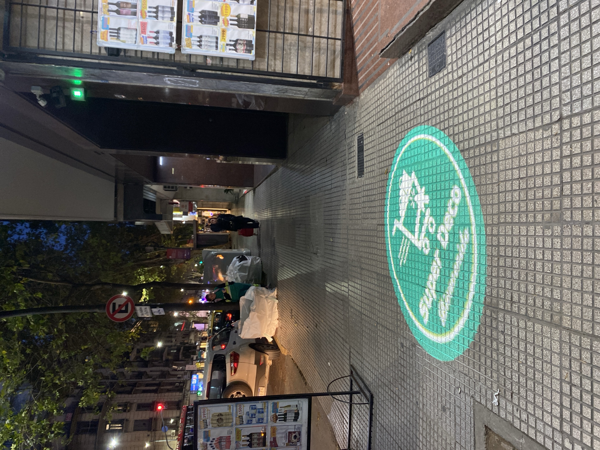 The vertically oriented image displays an urban street scene at night in Buenos Aires, Argentina. The perspective looks along a tiled sidewalk on the left of the frame, with a building wall running vertically on the right.
On the tiled building wall to the right, a circular green light projection illuminates a white graphic. This graphic depicts a stylized figure with a cane accompanied by a guide dog, encircled by text that reads "CUIDADO CON LA RAMPA" (Beware of the ramp), indicating an accessibility warning or feature. Higher up on this wall, a small, dark rectangular grate is visible.
To the left, the tiled sidewalk extends into the distance. In the foreground, an outdoor display stand showcases advertisements for bottled products, likely wine or spirits, with visible prices such as "3990," "1500," "6200," and the word "OFERTA" (offer). Above this, a building entrance with a metal gate also features similar advertisement posters displaying prices like "5600" and "2999." A small green light is visible near the top of this entrance, possibly a security indicator. Further down the sidewalk, piles of white garbage bags are visible, one of which is near a light-colored parked car. A person in a green uniform appears to be working near these bags, and another person in dark clothing with a red bag walks past.
Beyond the sidewalk, a street is visible with cars under artificial lighting. A street pole holds a red circular "no parking/stopping" sign, and an attached rectangular sign states "No estacionar de 7 a 21 hs" (No parking from 7 AM to 9 PM). Trees line the street, and distant buildings have illuminated windows, contributing to the city's evening ambiance.