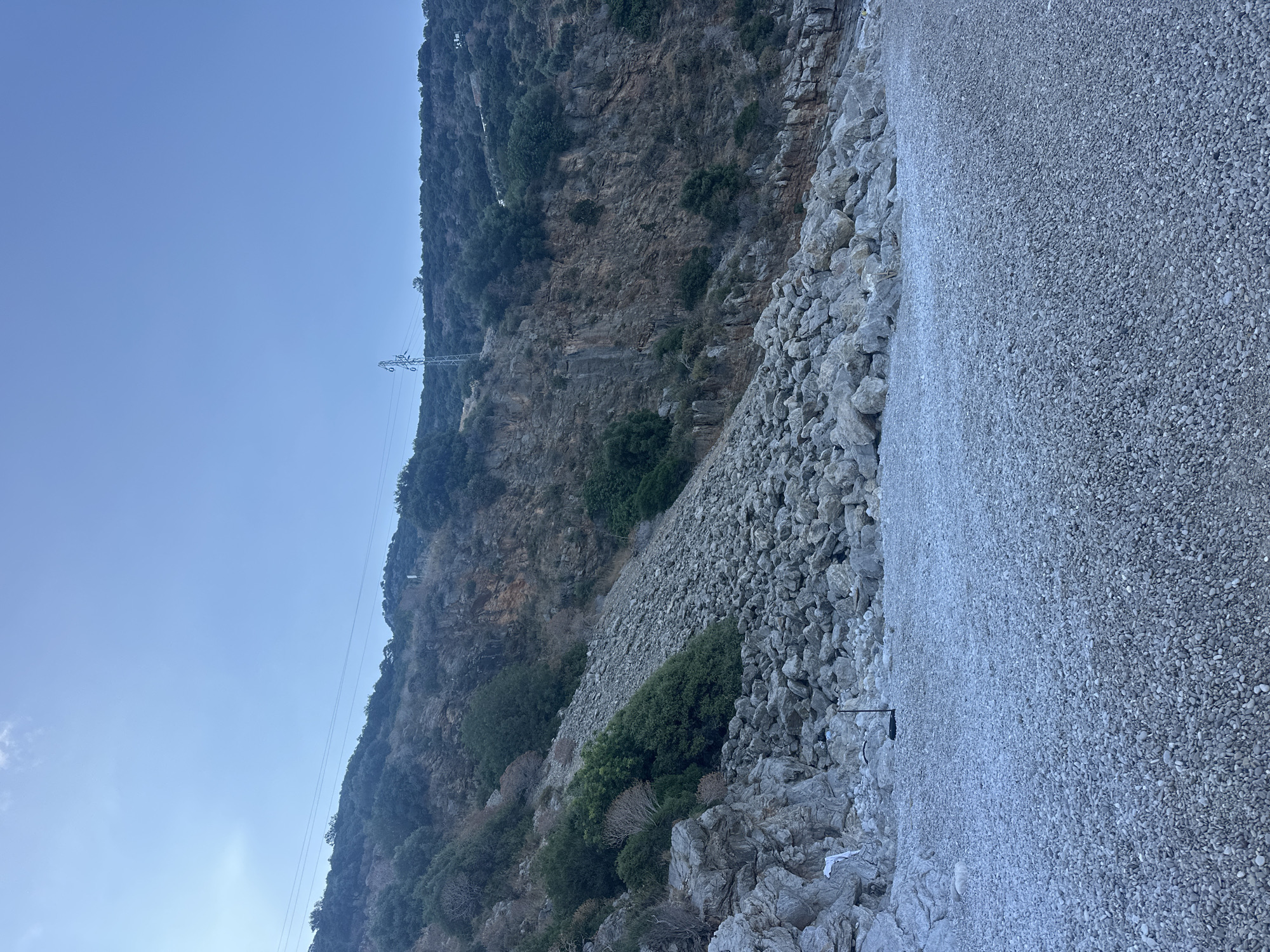 The image depicts a steep, vegetated cliff face meeting a pebble beach under a clear, light blue sky. The scene is located along the coast in the vicinity of Fethiye, Turkey, characteristic of the Mediterranean landscape with rocky terrain and coastal flora.
The upper portion of the image features a bright, mostly cloudless sky with a faint haze in the upper left quadrant. The mid-ground is dominated by a rugged, heavily vegetated mountain slope. Dense, dark green scrub and trees cover the upper and mid-sections of the slope, with exposed areas revealing reddish-brown and grey rock strata. A prominent scree slope, composed of numerous grey boulders and smaller rocks, descends from the mid-cliff to the beach. An electricity transmission tower (pylon) is visible on the upper slope towards the right, with power lines running horizontally across the mountain face. A white vertical marker is also present on the upper right cliff.
The foreground is a wide, flat pebble beach, uniformly covered in small, smooth, grey stones. Near the base of the cliff, the pebbles transition into larger, irregularly shaped boulders that appear to be part of the same geological formation as the cliff or detritus from it. No water is visible, but the presence of a beach implies proximity to the sea. On the lower left, among the larger rocks, a piece of white litter, possibly fabric or plastic, is visible. In the central foreground of the pebble beach, a dark, thin pole with a small, circular or disc-shaped top is inserted into the pebbles; its purpose is unclear. No individuals or animals are present in the scene. The lighting is soft and even, suggesting either early morning or late afternoon.