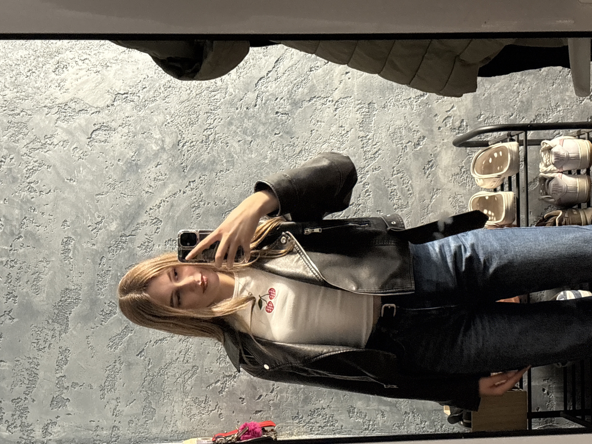 A young woman with blonde hair takes a selfie in a mirror. She wears a black leather jacket over a white shirt with a cherry design, and dark blue jeans. A coat rack with clothing and shoes sits to the right of the mirror. A fuzzy pink item is on a surface below the mirror, along with other small items. The wall behind the woman has a grey, textured finish. The location is not identifiable as Panagyurishte, Bulgaria.