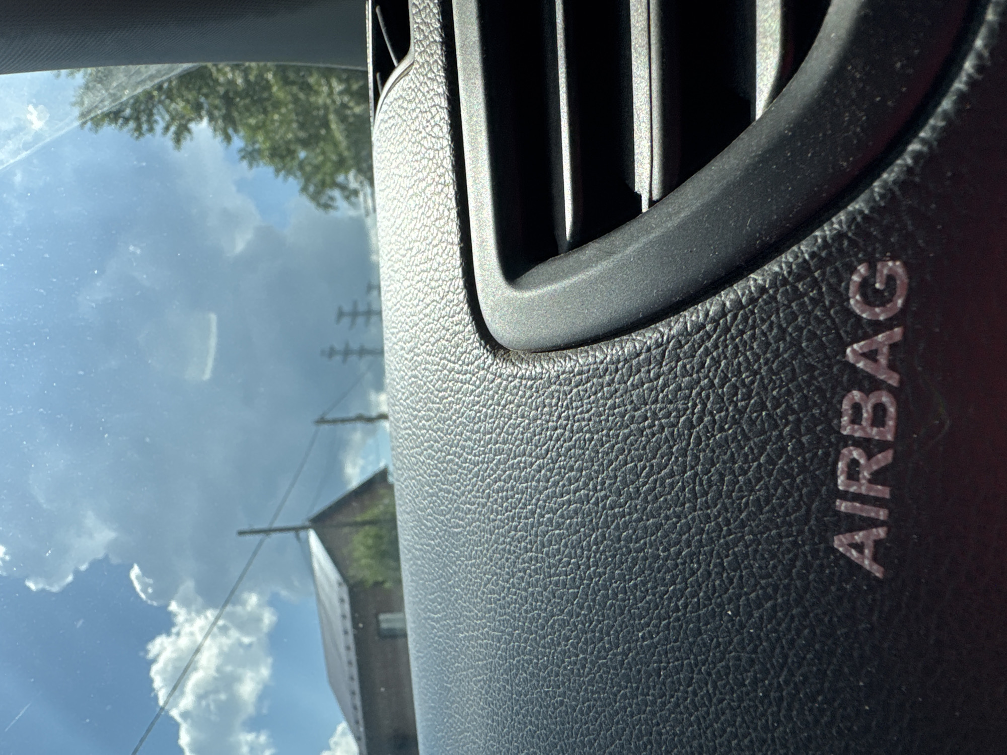 This is a close-up interior view from a vehicle, looking out through the windshield. The foreground is dominated by the textured surface of a car dashboard, with the embossed word "AIRBAG" visible in raised letters. To the right, a black air vent with horizontal slats is partially in frame. Beyond the dashboard, the scene through the windshield depicts a bright, partly cloudy sky with fluffy white clouds against a blue backdrop. In the middle ground, a dark-colored house with a steep roof and a chimney can be seen, situated behind power lines supported by utility poles. Green trees are visible to the right of the house, and further to the right, a larger tree with dense foliage is present. The overall impression is of a sunny day in a rural or suburban area. The location is specified as Chaikovsky, Russia.
