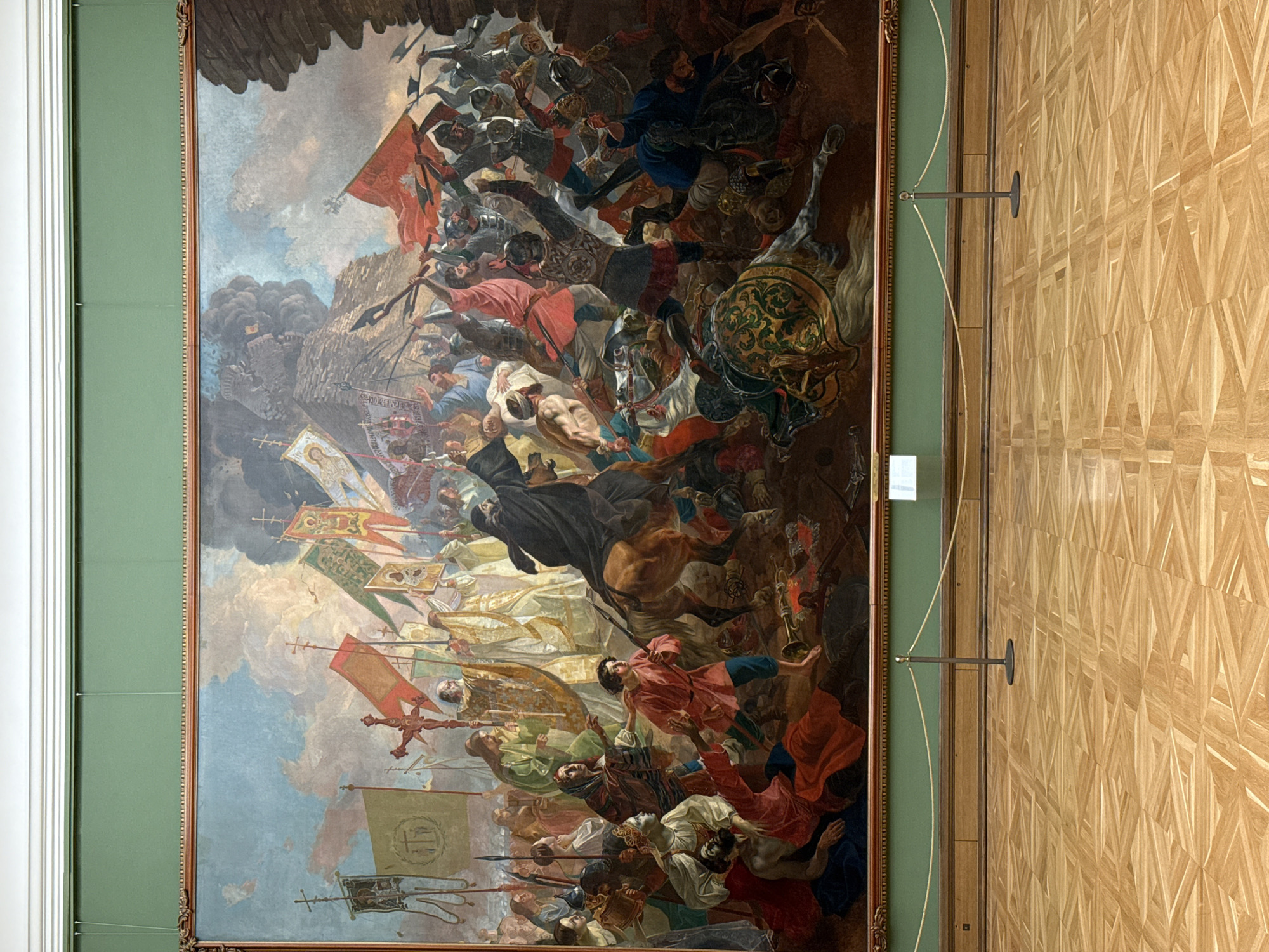 The image shows a large, vertically oriented painting depicting a historical battle scene. The painting is framed in an ornate, gilded frame. The battle scene includes numerous figures on horseback and on foot, engaged in combat. Flags and banners are visible amidst the chaos. The color palette is dominated by reds, blues, browns, and creams, giving the painting a dynamic and dramatic feel. The painting is displayed on a wall, likely in a museum or art gallery.
To the right of the painting is a light-colored wooden wall with a geometric pattern. A metal stanchion or barrier is attached to the wall, presumably to prevent visitors from getting too close to the artwork. There is also a light green wall to the left of the painting. The painting is located in Moscow, Russia.