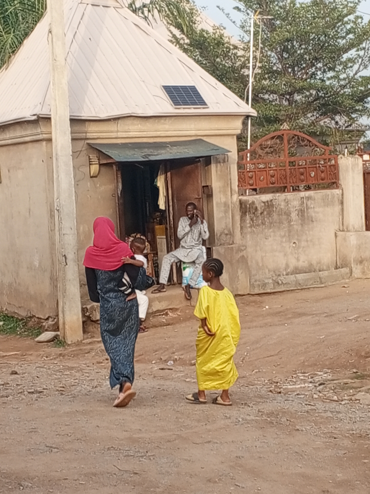 The image shows a street scene in an urban area. In the foreground, a woman wearing a dark blue patterned dress and a red hijab is carrying a small child. A girl wearing a bright yellow dress walks to her right. In the background, a man in patterned clothing is sitting in a chair in front of a small building with a grey roof and a solar panel. He appears to be using a mobile phone. Behind the building is a fence with an ornate metal gate. The ground is unpaved.