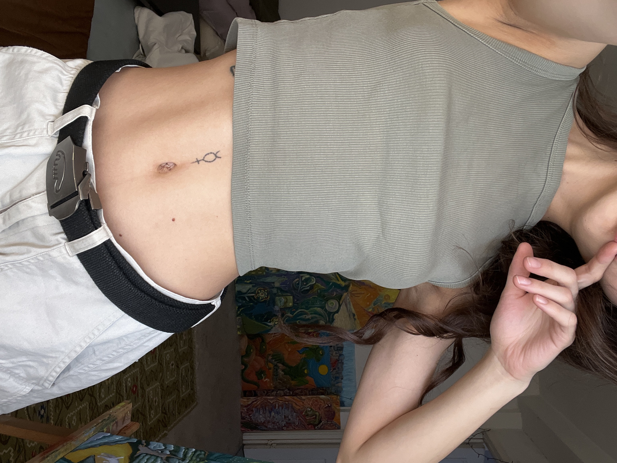 A woman is visible, wearing a light green tank top and white pants with a black belt. A tattoo of a symbol is visible on her stomach, near her navel. She is partially in the foreground. Part of a painting is visible in the background.
The location in Krasnoyarsk, Russia cannot be determined from the image.