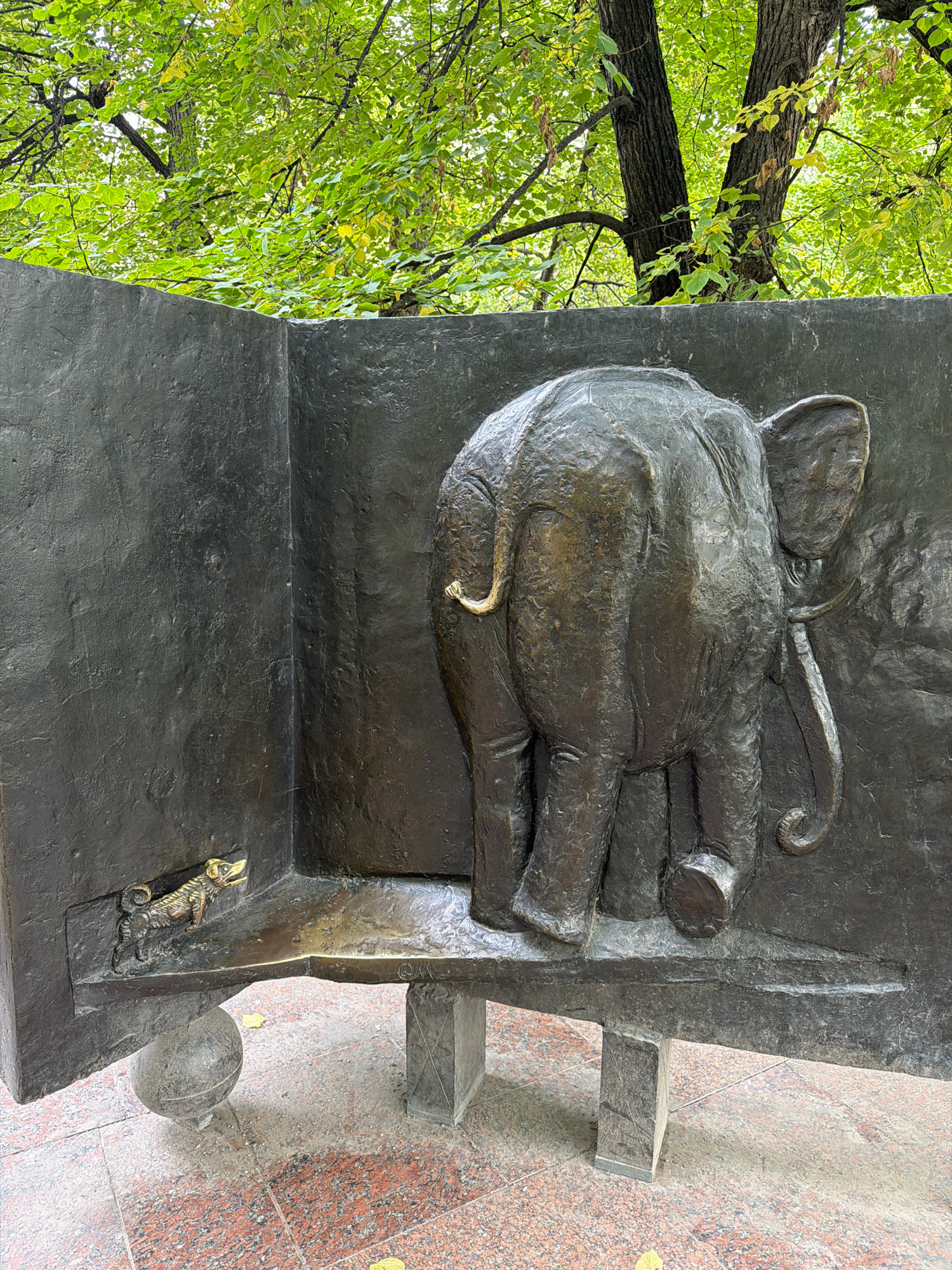 This image shows a bronze sculpture on a stone pedestal, situated outdoors under a canopy of green trees with dappled sunlight. The sculpture depicts an elephant's rear end, facing away from the viewer, and a small dog figure in the lower left corner. The elephant is rendered in relief on what appears to be an open book or a wall-like structure. The dog is a small, detailed metal casting, positioned on a shelf-like platform. The sculpture is dark bronze, with some lighter highlights from the lighting. The pedestal is made of speckled granite. The ground around the pedestal is paved with reddish-pink granite tiles. The trees overhead have green leaves, some tinged with yellow, suggesting it might be early autumn.
This sculpture is part of the monument to Mikhail Bulgakov, located at 35 Bolshaya Sadovaya Street in Moscow, Russia. It is situated in the courtyard of the Bulgakov Museum, which is housed in the apartment where Bulgakov lived and wrote "The Master and Margarita." The elephant and dog are characters or elements from Bulgakov's literary works.