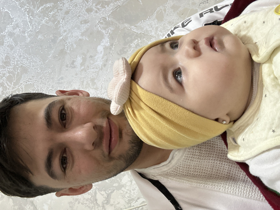A close-up captures a man and a baby. The man, with dark hair and a trimmed beard, is positioned to the left. The baby, wearing a yellow headband with a cream-colored bow and a light yellow outfit with stars, is to the right. The baby's eyes are looking upwards. Both are resting on a surface with a white patterned background. A white mesh shirt and black strap are also visible.