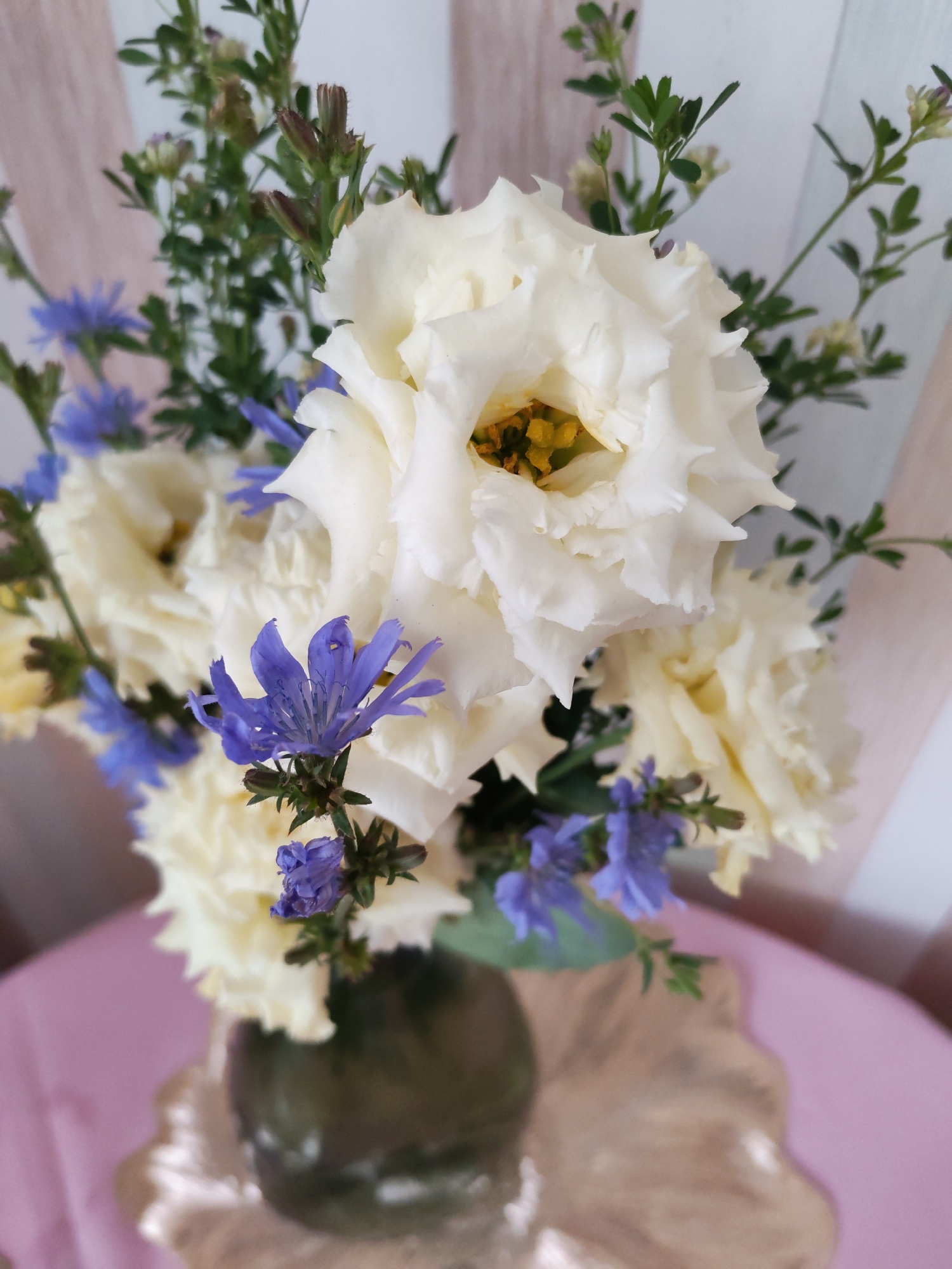 A bouquet of flowers, featuring large white roses and small blue wildflowers, is arranged in a dark green vase. The bouquet is placed on a pink surface, which appears to be a tablecloth, and rests on a decorative gold platter. The background consists of vertical light-colored stripes, suggesting a wall or screen. The image was taken indoors, likely in a domestic setting. The specific location within Izhevsk, Russia, cannot be determined from the visual information provided.