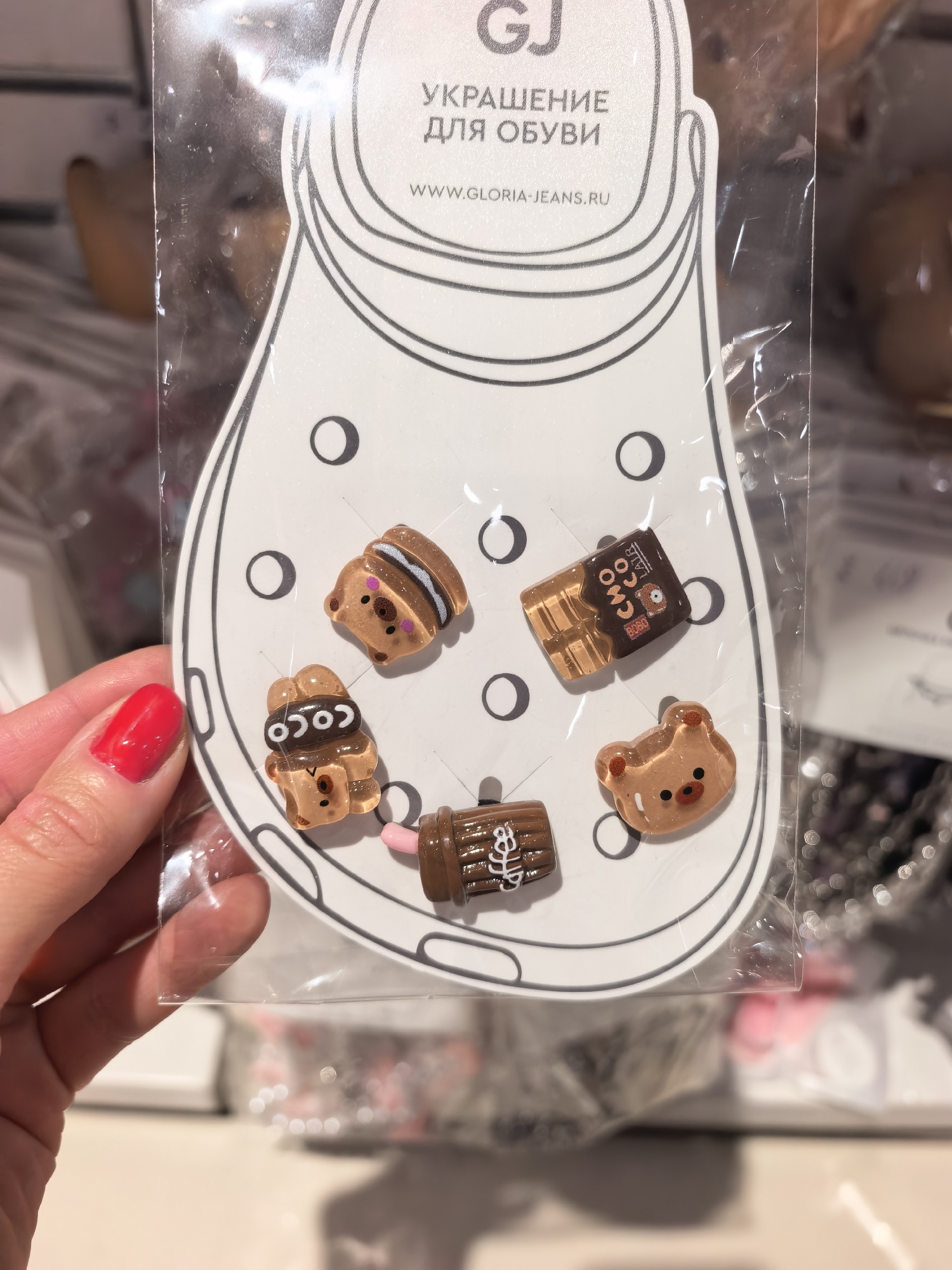 A hand with bright red nail polish is holding up a package of five small, decorative charms for shoes. The charms are shaped like various food items and animals, including a bear face, a chocolate bar, a cookie sandwich, a brown animal wearing a chocolate-colored hat with "COCO" written on it, and a brown cup with a pink straw and white frosting. The package is designed to look like the outline of a shoe. Text on the package reads "GJ", "УКРАШЕНИЕ ДЛЯ ОБУВИ", and "WWW.GLORIA-JEANS.RU". The background is blurred, but appears to be an indoor retail environment with shelves and other merchandise. There is no specific indication of the location being Sterlitamak, Russia.
