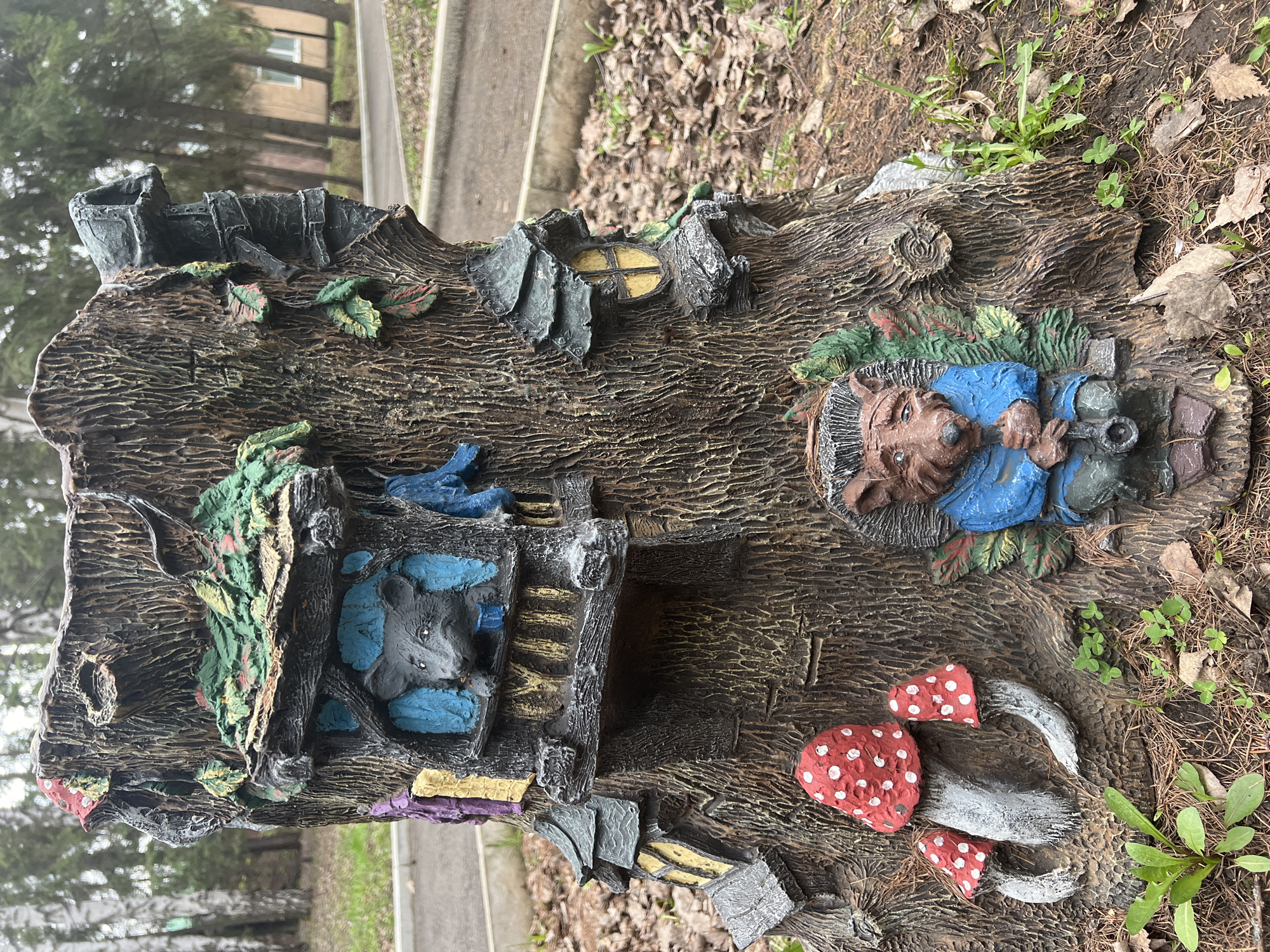 The image shows a decorative tree stump, potentially located in Sanatoriya Yumatovo Imeni 15-Letiya Bassr, Russia (as per provided location). The stump features various molded and painted elements, including a sleeping figure resembling a hedgehog or similar creature wearing a blue shirt, a mouse-like figure within a dwelling, red-capped mushrooms, and a small window. The environment surrounding the stump consists of dirt, leaves, and some vegetation, with a paved path and building visible in the blurred background. The photograph is taken at a relatively close range, providing a detailed view of the stump's textured surface and painted components.