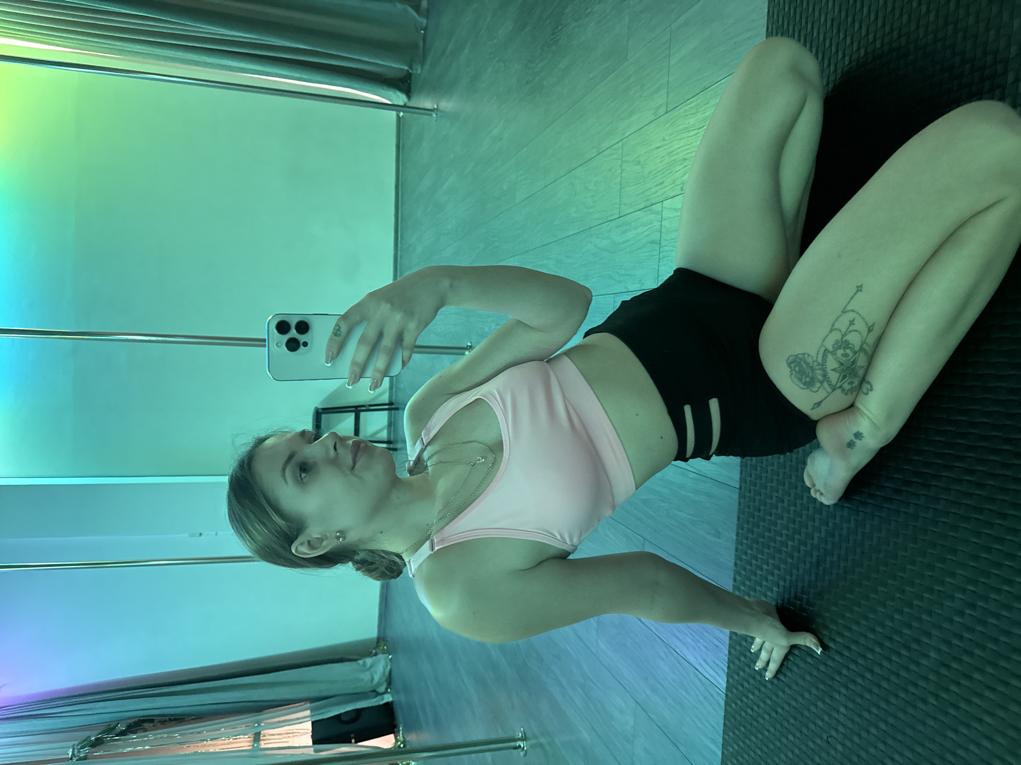 A woman is taking a selfie while sitting on a black yoga mat indoors, possibly in a studio or fitness center in Batumi, Georgia. The woman is fair-skinned, wearing a light pink sports bra and black shorts with a cutout design on the sides. She is holding a white smartphone in her right hand to capture the image. Her hair is pulled back, and she is wearing jewelry, including earrings and a necklace. A tattoo is visible on her right leg. The room is illuminated with a vibrant blue-green light, possibly from LED panels or reflective surfaces. A chrome pole structure, likely part of a fitness or dance setup, is seen in the background along with gray blinds. The background suggests the location could be in a modern building or converted space.