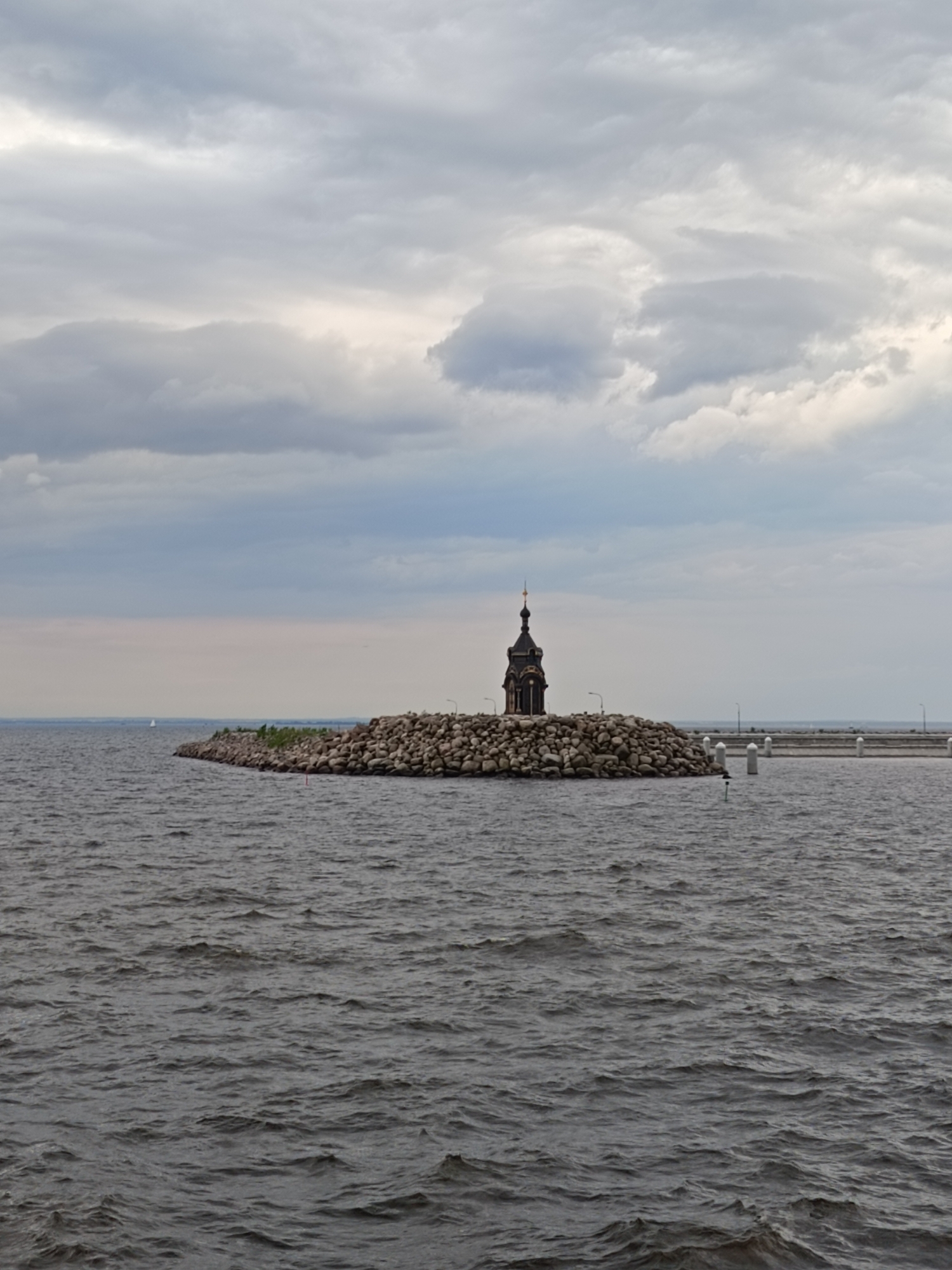 The image displays an overcast marine landscape, centered on a dark, ornate chapel situated on an artificial rocky island. The scene depicts the Lighthouse Chapel of St. Nicholas in Kronstadt, an island city within Saint Petersburg, Russia.
The sky occupies the upper portion, characterized by a uniform expanse of muted gray and white clouds, indicating an overcast condition with no direct sunlight. The clouds show varying densities, with some brighter patches.
The lower two-thirds of the image consist of a large body of dark gray water. The water surface is textured with numerous small, choppy waves, suggesting movement or wind.
In the middle ground, an artificial island, or breakwater, is constructed from a dense collection of large, rounded, gray boulders and rocks. Sparse green vegetation is visible on parts of the rocky embankment. Several streetlights are positioned along the top of this rocky structure.
The central subject is a dark-colored, multi-tiered building with a prominent spire, resembling a chapel or a lighthouse with a chapel design. The spire is topped with a distinct, possibly golden or bronze, finial. The building's main body appears dark, possibly black or very dark brown, with architectural details indistinct due to distance and lighting.
To the right of the artificial island, a lighter-colored pier or additional breakwater extends horizontally across the frame. This structure features several evenly spaced, thick, white vertical posts along its edge. In the water near the island and pier, a green cylindrical navigational buoy is visible, along with another white post or buoy.
In the far distance on the left horizon, a small white sailboat with a mast and sail is visible. No other identifiable people or animals are present in the scene. The overall color palette is dominated by cool grays and dark tones, reflecting the overcast weather.