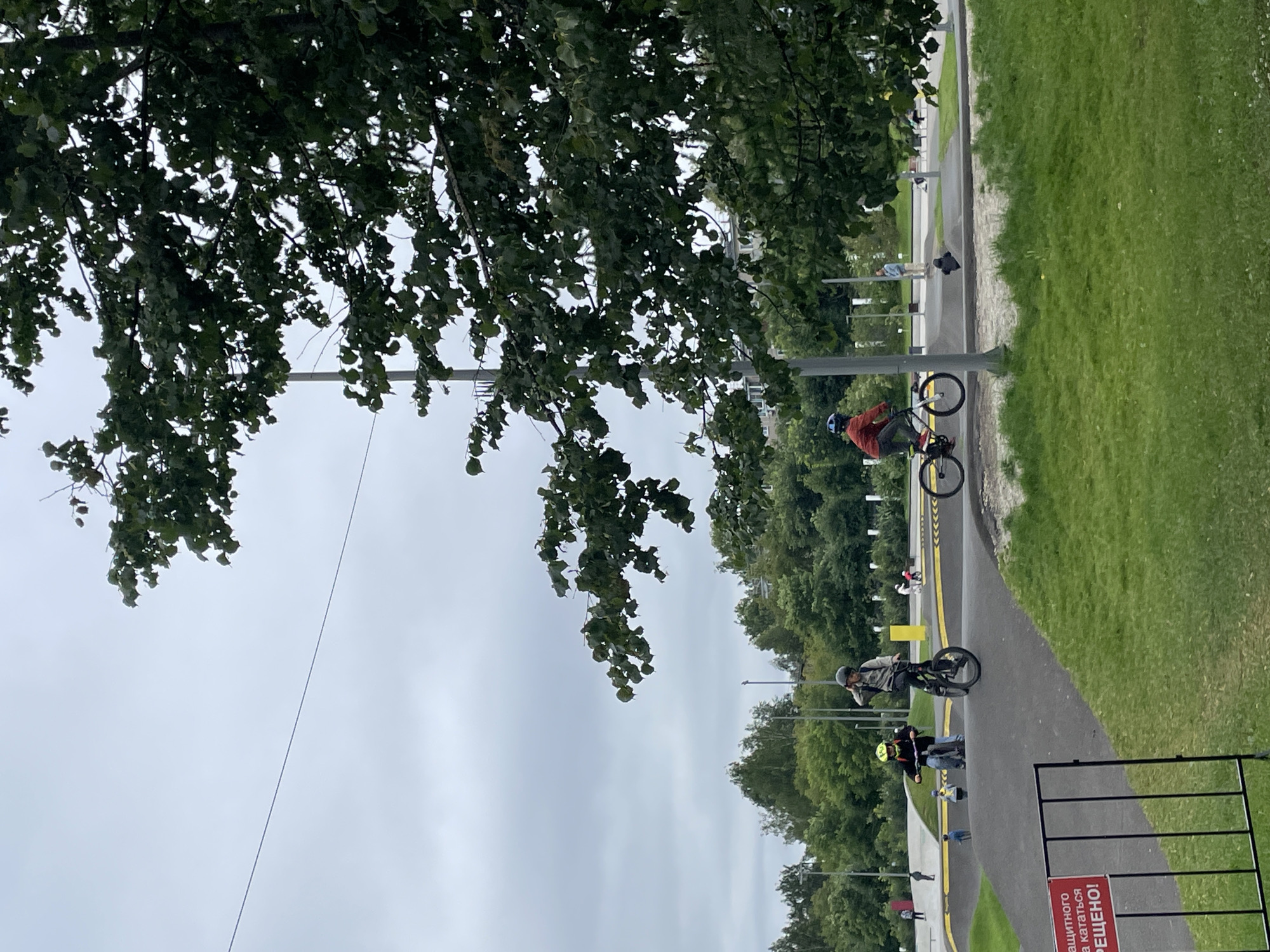 The image depicts a bicycle pump track with several individuals engaged in cycling activities. In the foreground, a red and white sign with Russian text is partially visible, indicating a prohibition. A black metal fence borders a grassy area.
In the middle ground, two young people are on bicycles. The person on the left, wearing a yellow helmet and a dark jacket, is on a smaller bicycle. The person on the right, wearing a light-colored helmet and a grey jacket, is on a larger bicycle with a single wheel visible, appearing to be performing a trick. Further to the right, another individual in an orange jacket is riding a bicycle.
The background features a tree-lined area with several light poles. The sky is overcast, contributing to a muted lighting condition. The pump track itself is characterized by a dark, asphalt-like surface with undulating terrain designed for cycling. The overall environment suggests an outdoor recreational park.
This scene is located in the city of Kirov, Russia, specifically at the Kirov Bicycle Pump Track located in the Zarechny District.