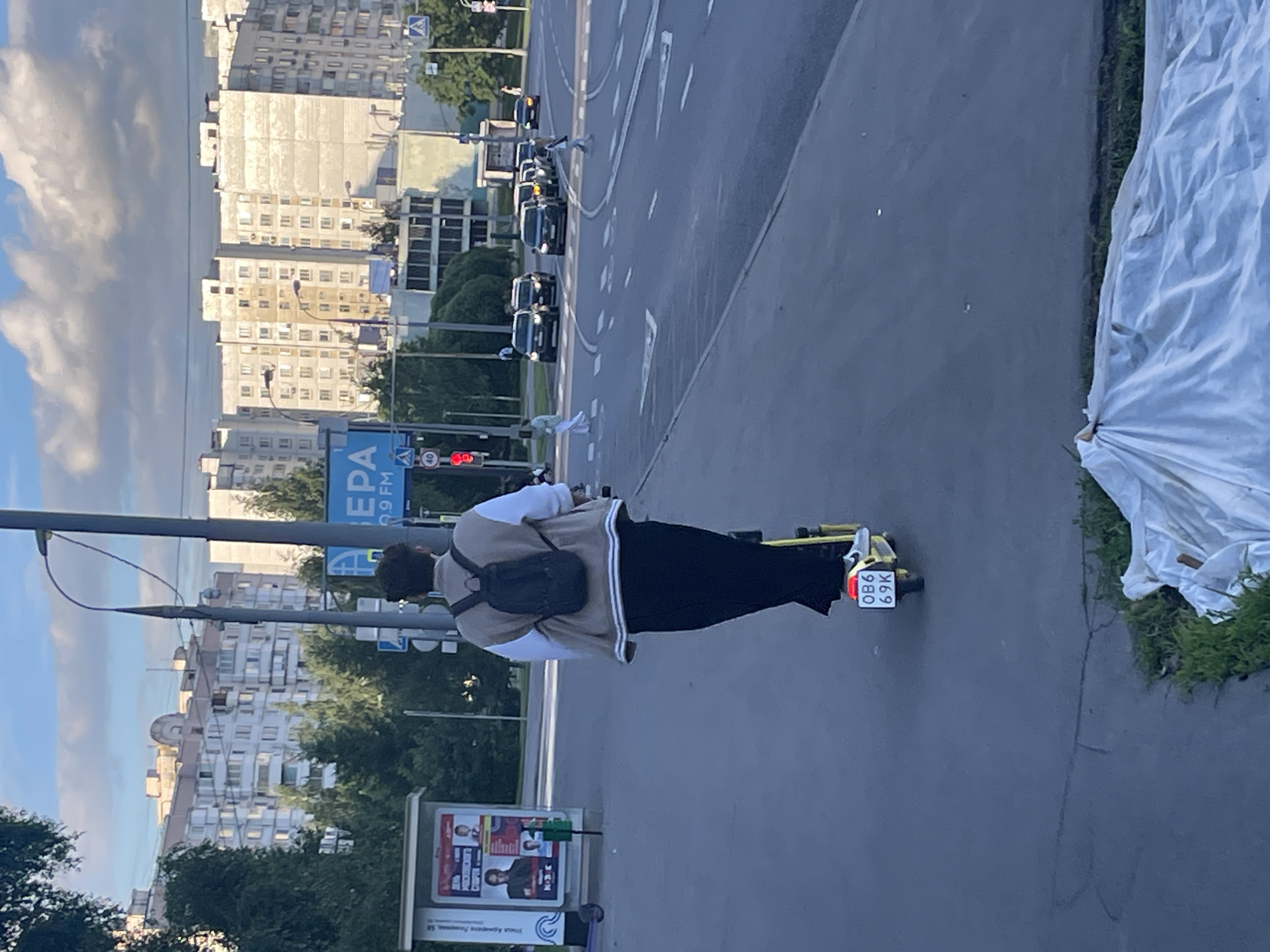 The image shows a person riding an electric scooter on a city street in Moscow, Russia. The person is wearing a light-colored sweater with white stripes and dark pants, and has a black backpack. The scooter has a license plate that reads "0B6 69K". In the background, there are multi-story apartment buildings, trees, and a street with cars. A traffic light is visible, and it appears to be red. The sky is cloudy, suggesting it might be daytime, possibly late afternoon. The scene takes place on a paved road, likely a sidewalk or bike lane next to a main road. There is a large advertising billboard to the left of the scooter rider. The overall mood seems to be casual and urban, with the rider enjoying a leisurely ride through the city.