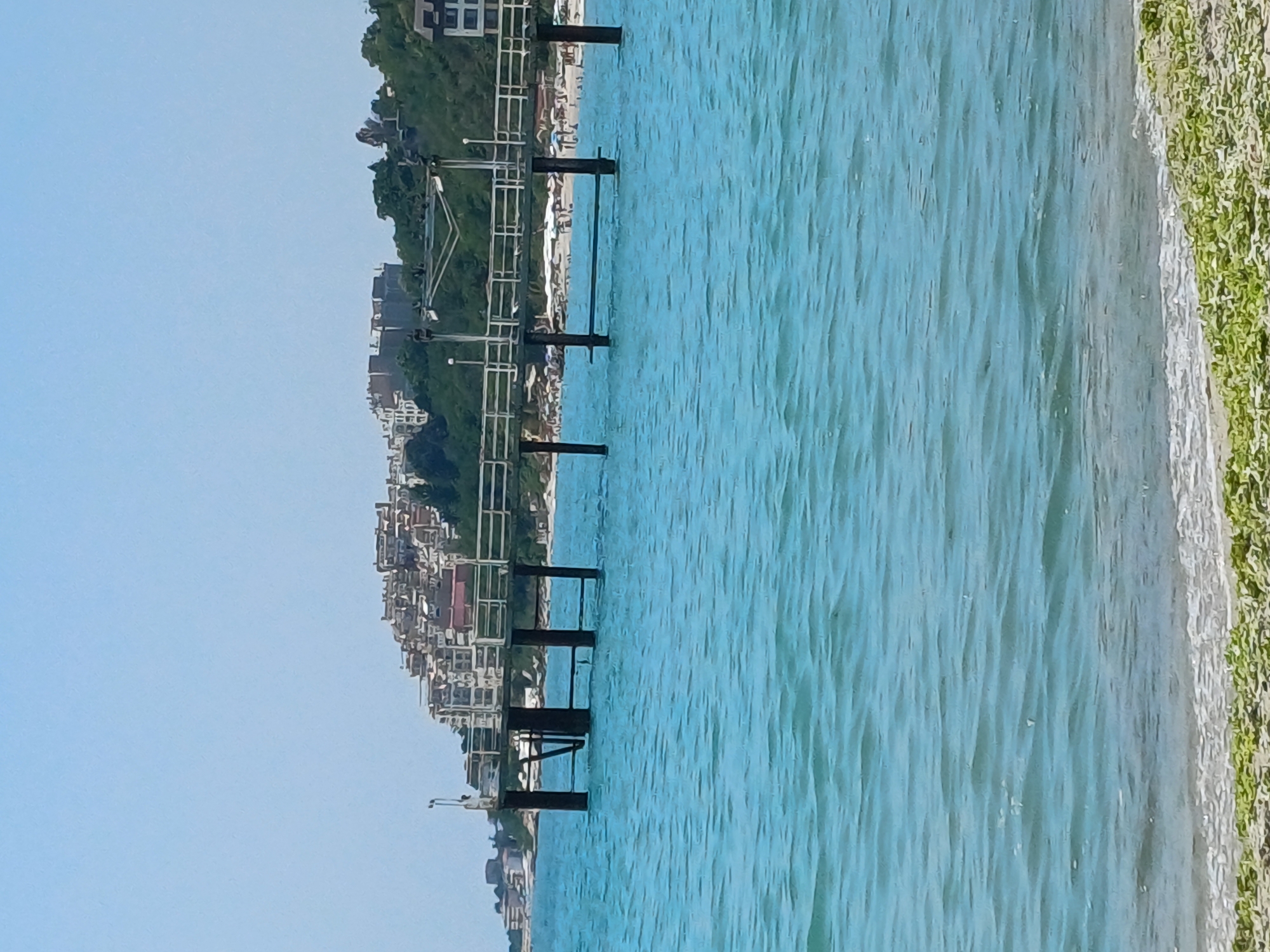 The image captures a view of the Black Sea from a beach in Varna, Bulgaria. In the foreground, the turquoise water displays gentle ripples and a line of green seaweed along the sandy shore. A wooden pier extends into the sea, supported by dark pilings. Beyond the pier, a bustling beach scene unfolds with numerous beach umbrellas, lounge chairs, and people. In the background, a hillside is densely populated with white buildings, suggesting a resort town. The sky is clear and blue, indicating fair weather.