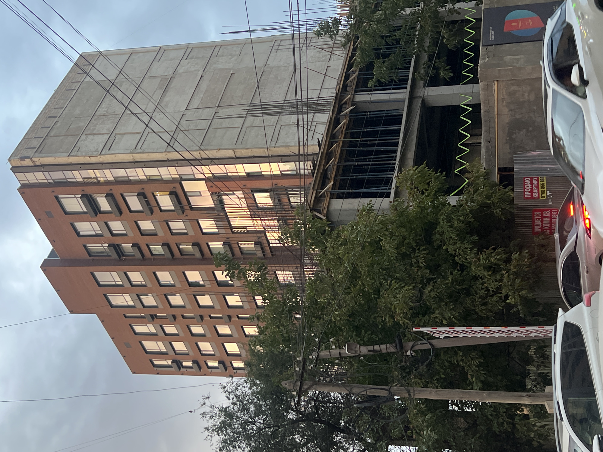 The image shows a tall, multi-story building under construction in Bishkek, Kyrgyzstan. The building features a facade of red brick on one side with numerous windows reflecting the sky. The adjacent side of the building is unfinished concrete with visible rebar. At the lower levels of the building, there are visible construction elements like scaffolding and concrete supports. Surrounding the building, there are lush green trees, utility poles with electrical wires, and a street with cars. A red and white striped barrier is visible near the utility poles. In the foreground, several cars are parked or driving on the street. Signs are posted on a fence in front of the construction site, one of which reads "ПРОДАЮ КВАРТИРУ" (Apartment for Sale). The sky is overcast.