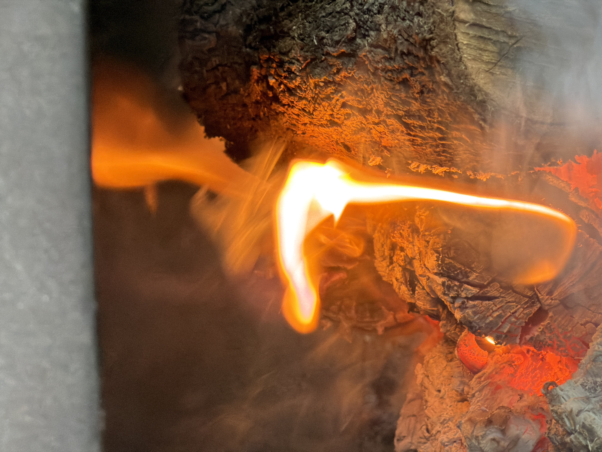 A close-up shot captures flames engulfing wood inside a fireplace. The fire is intense with bright orange and white colors dominating the scene. Dark charred wood is present, contrasting with the vibrant flames and faint smoke rising. No people or specific actions are visible, the focus is on the fire itself. It's not possible to determine if this scene is located in Hout Bay, South Africa based solely on the image.