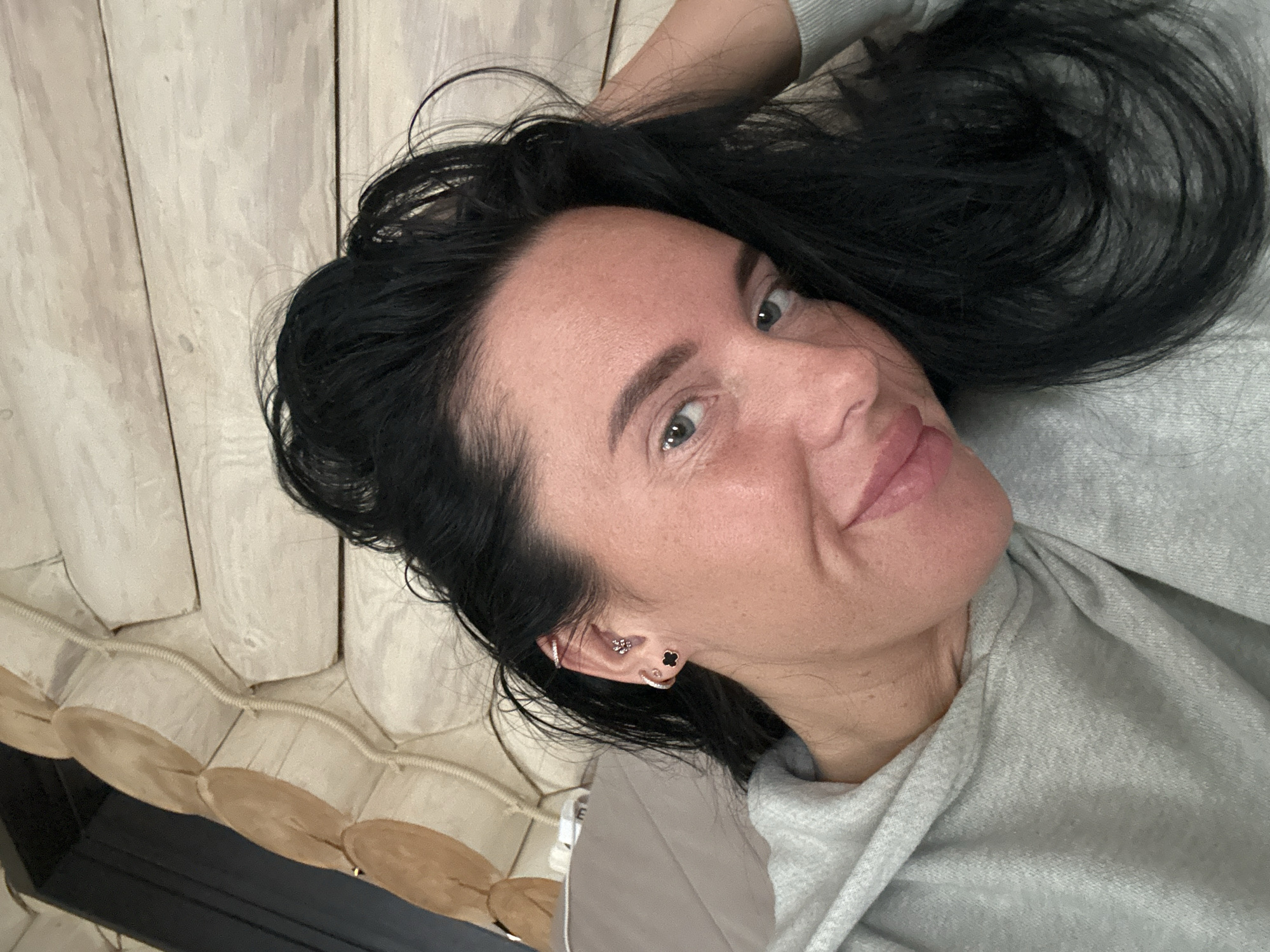 A woman with dark hair and light eyes is lying on a bed, wearing a gray sweatshirt. She is facing the camera with a slight smile. She has multiple earrings in her left ear. The bed has a wooden headboard and a decorative row of connected, round, tan and dark brown elements along the edge of the bed frame. The photo appears to be taken indoors. It is unknown if this photo was taken in the city of Ольховка, Russia.