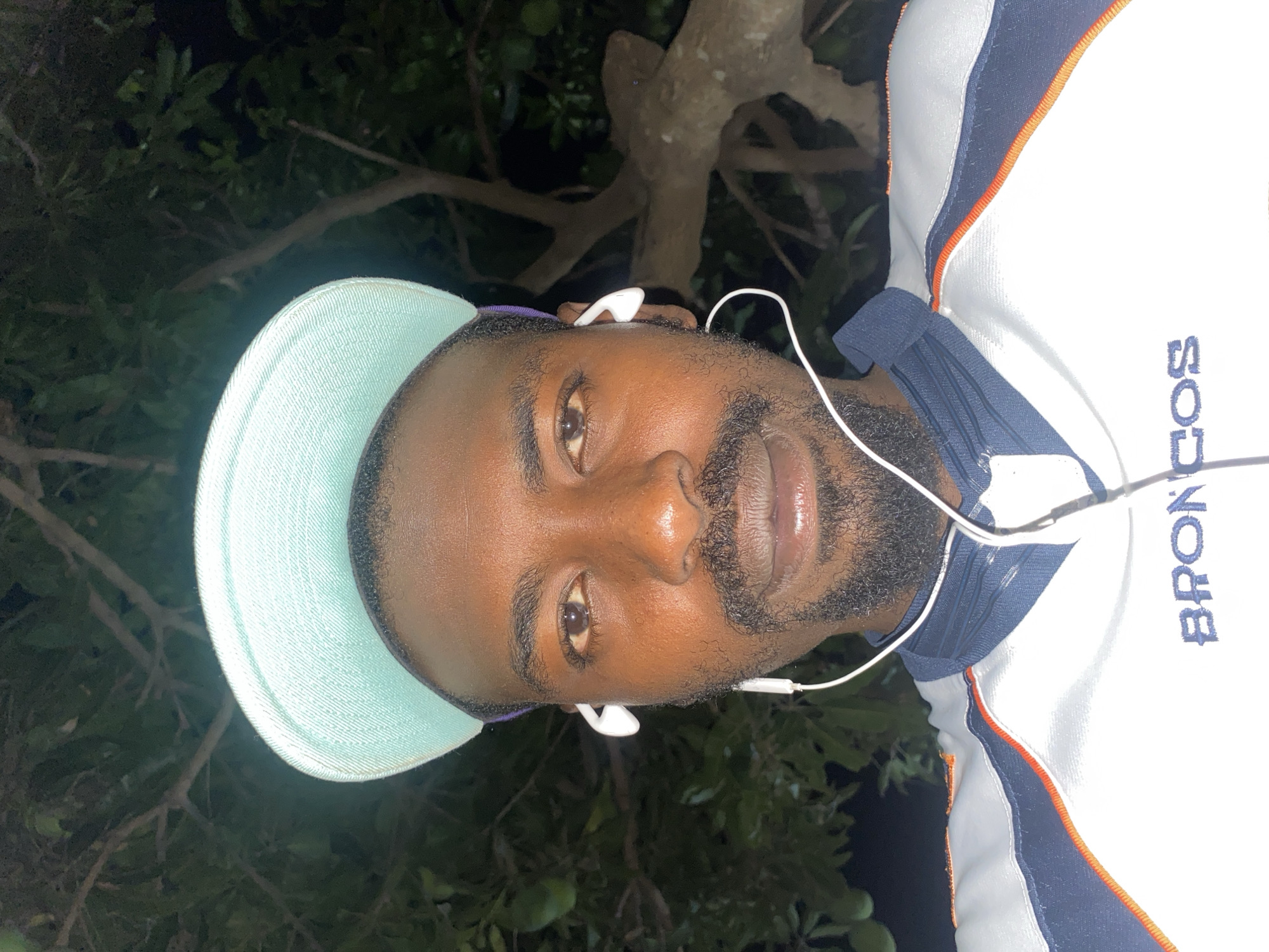 A portrait-style image, rotated ninety degrees counter-clockwise, features a man with dark skin and a beard, looking directly at the camera. He has short hair and is wearing a light blue baseball cap. White wired earbuds are visible in his ears, with the wires draped downwards. The man is wearing a white collared shirt with blue shoulders and orange piping. The word "BRONCOS" is distinctly visible in blue lettering on the white part of his shirt, near his left shoulder.
The background is dark, indicating nighttime or a dimly lit outdoor setting. Dark green foliage and tree branches are discernible behind him, suggesting he is outdoors, possibly beneath trees. The man's expression is neutral to slightly contemplative. The image captures a casual moment, possibly a selfie or portrait, with artificial light illuminating his face against the natural, shadowed backdrop. The setting is consistent with an outdoor location in Jos, Nigeria.