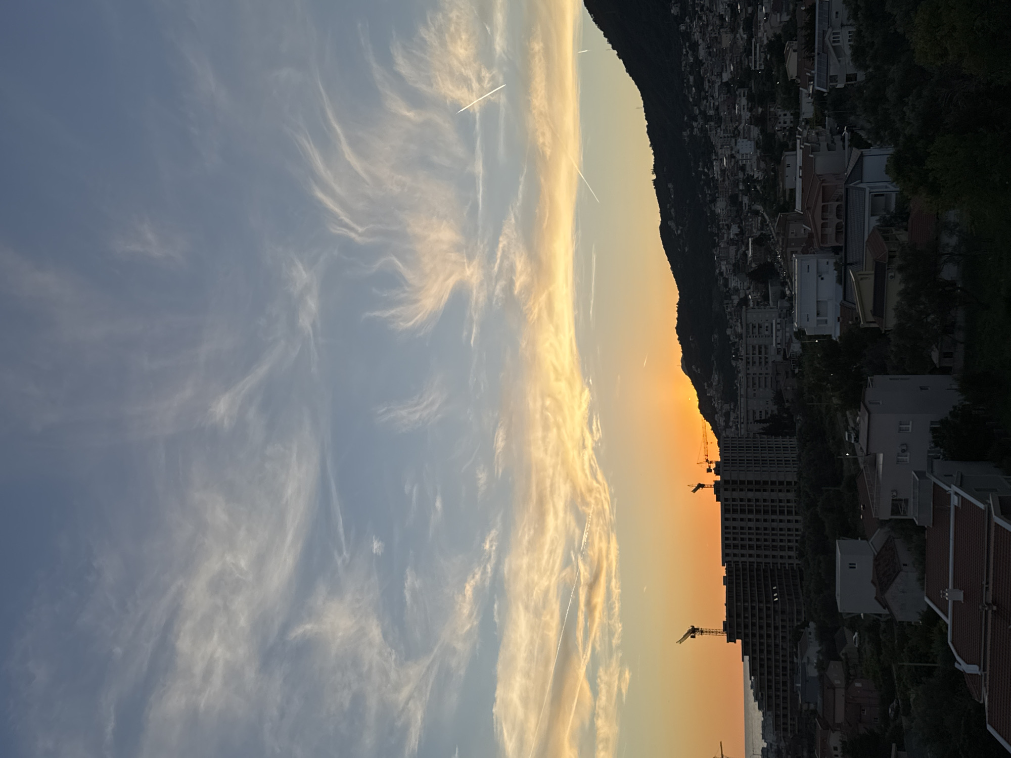 The image depicts a city skyline at sunset in Šušanj, Montenegro. The sky is a gradient of blue, with wispy white clouds and orange hues near the horizon, indicating the setting sun. Several contrails from aircraft are visible in the sky. In the foreground, a large, unfinished building with cranes on top is under construction, likely a hotel or residential complex, situated near the coast. Beyond this construction, a dense urban area with numerous houses and buildings stretches across a hillside, leading up to a dark, mountainous backdrop. Trees and vegetation are scattered throughout the urban landscape. The overall scene captures a developing coastal town during the golden hour. There are no people visible in the image.