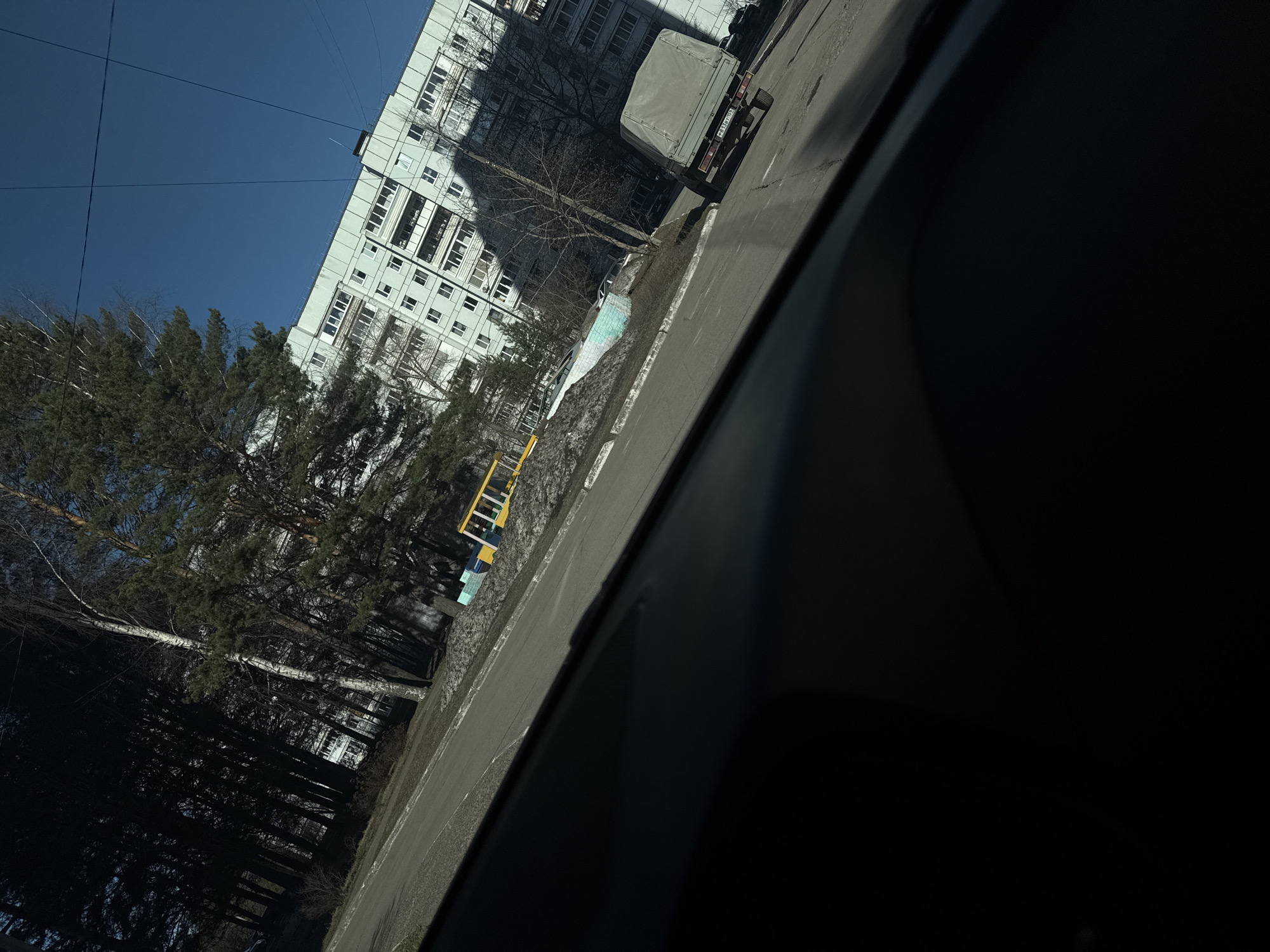 The image shows a street scene in what appears to be a residential area. A multi-story building with numerous windows is visible in the background. A truck is parked on the side of the road. Trees are present, some of which are bare and others with green foliage. A yellow object (possibly a piece of street furniture) is also present on the side of the road. The image seems to be taken from inside a vehicle due to the presence of what is likely part of a car door or window frame.
The location is Chaikovsky, Russia.