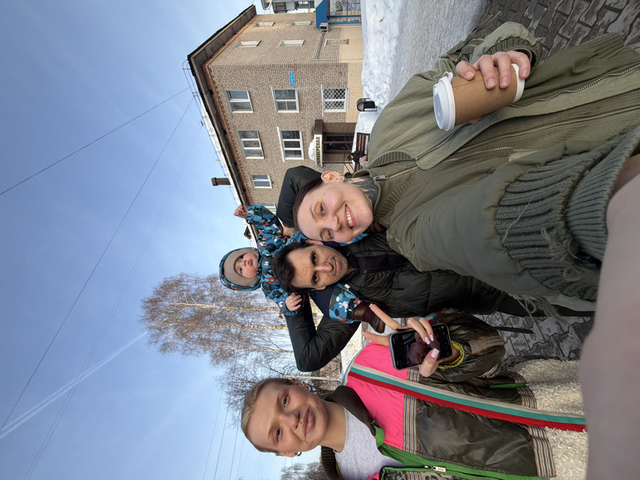 A group of four individuals, comprising three adults and one child, is depicted in a selfie taken outdoors on a clear, sunny winter day in Chaikovsky, Russia. The individual taking the photograph, a woman with dark hair and visible neck tattoos, is on the right, smiling and holding a disposable coffee cup in her right hand. To her left, a man with dark hair and a somber expression wears a dark puffer jacket and supports a small child on his shoulders. The child, dressed in a blue and brown patterned winter suit and a beige knit hat, looks forward with its right arm raised. To the man's left, a woman with blonde hair, wearing a multi-colored jacket (pink, green, brown, white), holds a dark smartphone with a pink case, displaying a blurred image on its screen, and offers a slight smile.
The scene features snow covering the ground beside a paved area in the foreground. In the background, a three-story, light-colored brick building with multiple windows is visible. A sign above its entrance reads "ПАРИКМАХЕРСКАЯ" (HAIRDRESSER). Bare deciduous trees and overhead power lines are visible against a bright blue sky, which also shows a distinct airplane contrail. A trash bin and a bench are situated near the building's entrance, and a partial blue structure is visible on the far right.