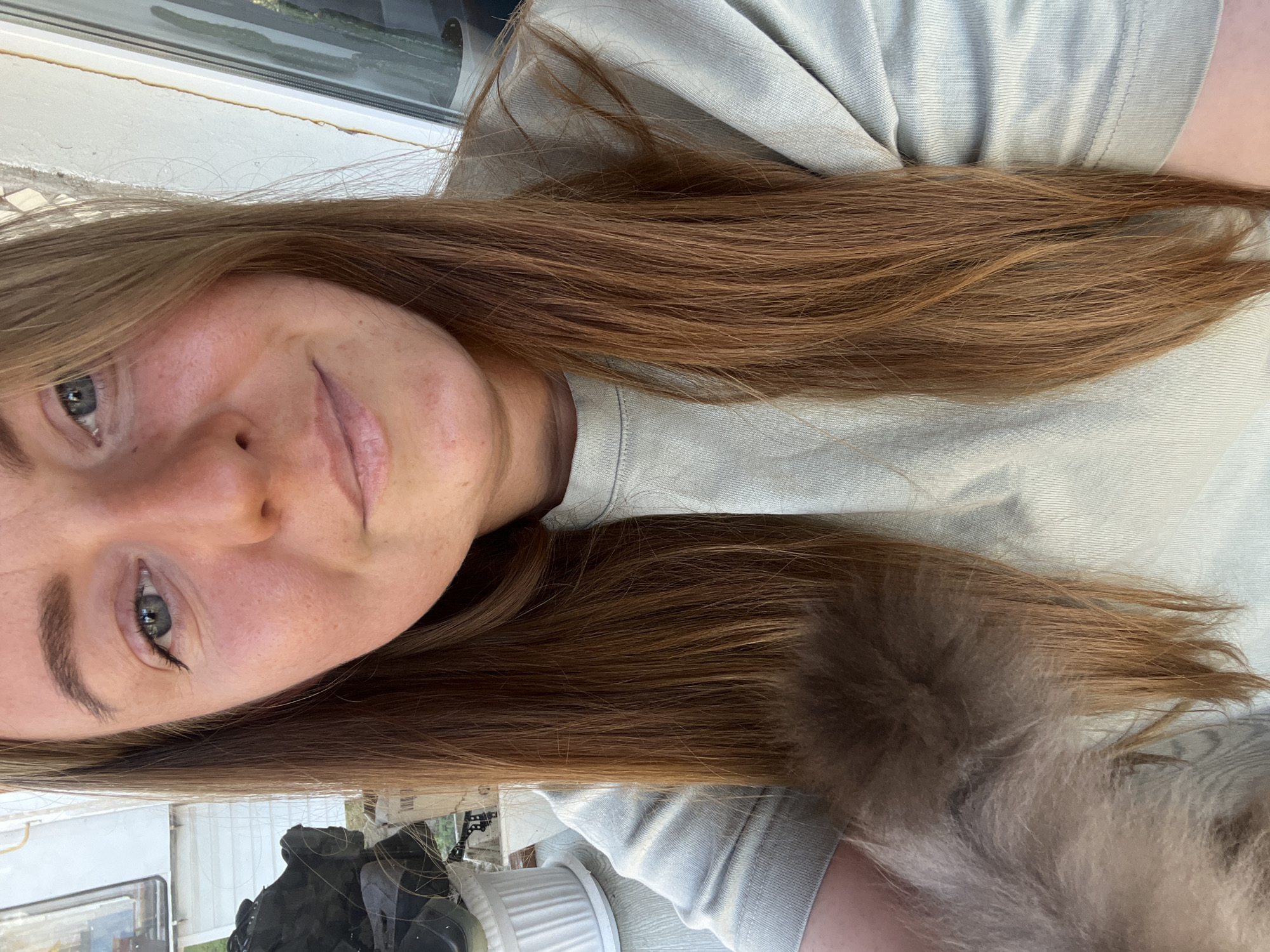 A young fair-skinned woman with long brown hair is centered in the frame. She has light-colored eyes and a slight smile. She is wearing a light-colored t-shirt. Part of what appears to be a cat's fluffy fur is visible in the lower right corner of the image. There is a window behind her and some indistinct objects are visible through it. There is no evidence in the photo to indicate that it was taken in Razdolye, Russia.
