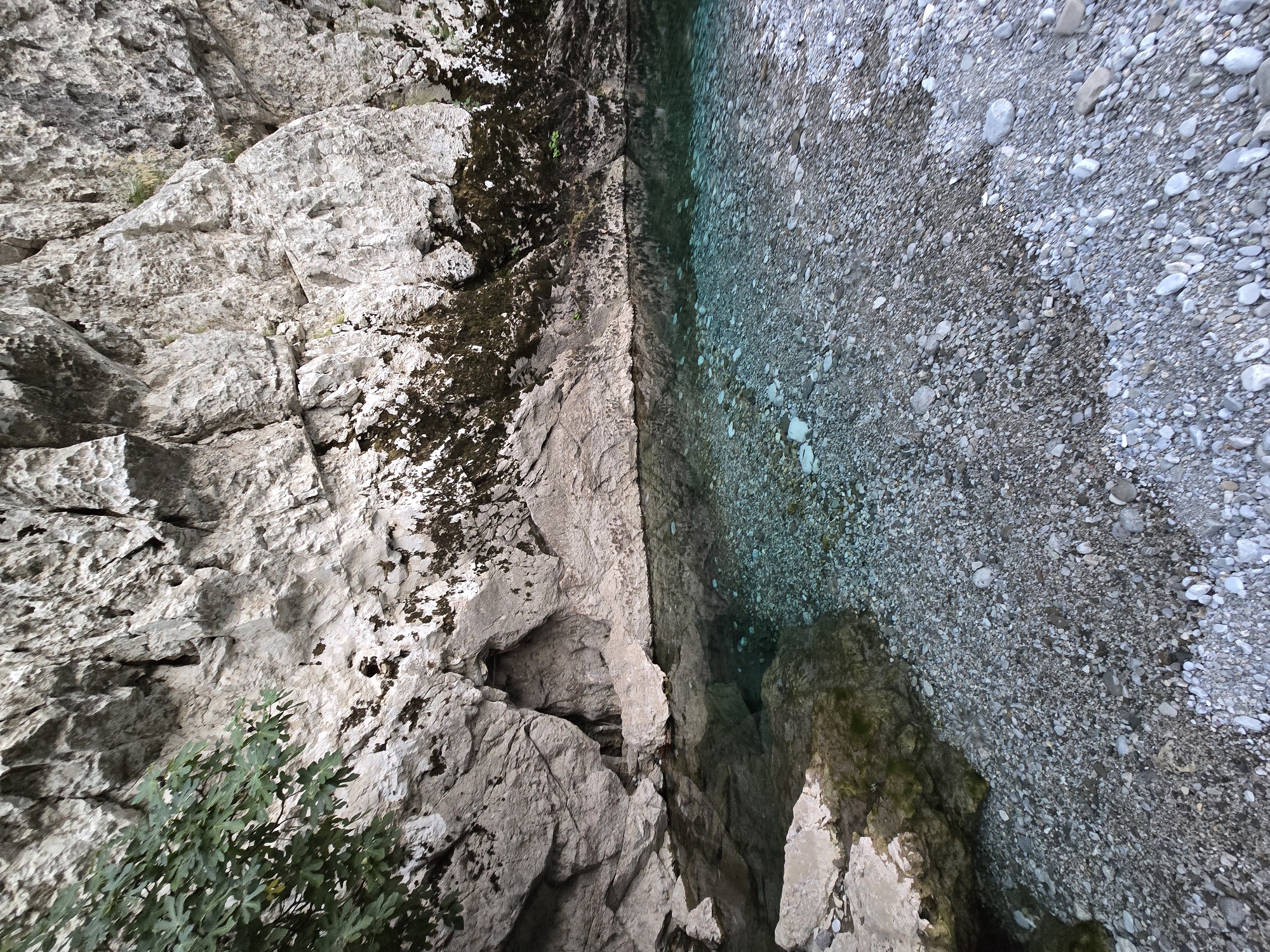 The image depicts a clear, shallow body of water with a rocky bed, situated at the base of a large, textured rock face. The water is a striking turquoise color, with the rocky bottom visible through its transparency. The rock face is predominantly light gray, with dark patches of moss and vegetation clinging to its surface. On the left side of the image, a branch with green leaves overhangs the water. The foreground consists of a stony shoreline, composed of numerous small, smooth pebbles in varying shades of gray and white. The scene appears to be a natural outdoor environment, possibly a river or a secluded pool within a rocky gorge, located in Bioče, Montenegro. There are no people or visible signs of human activity in the image, suggesting a remote and untouched natural setting. The lighting suggests it is daytime, with soft light illuminating the scene.