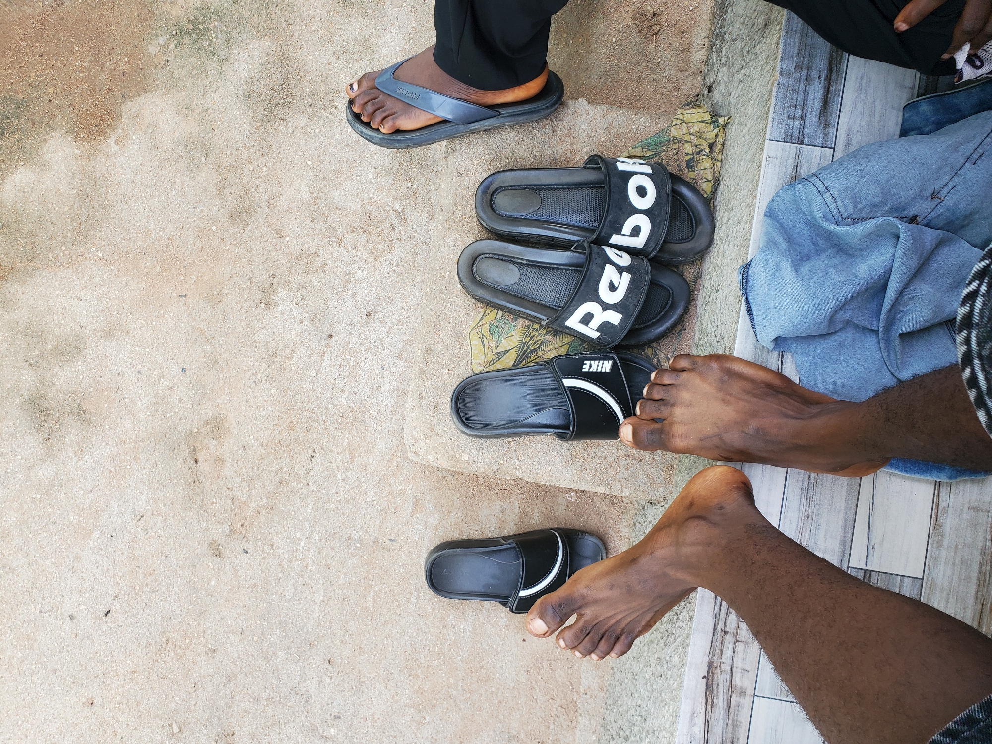 In Jalingo, Nigeria, this image captures a close-up overhead view of several pairs of open-toed sandals and bare feet on a textured, light-colored concrete ground. Two pairs of black Nike slides are positioned on the left, with the left foot of an individual resting near one of them. To the right, a pair of black Reebok slides are placed together, and a person's right foot, wearing a gray flip-flop, is visible near them. A folded blue denim garment lies on the ground between the two sets of footwear, partially obscuring the concrete and a patterned rug underneath. The overall impression is one of casual relaxation in an outdoor setting.