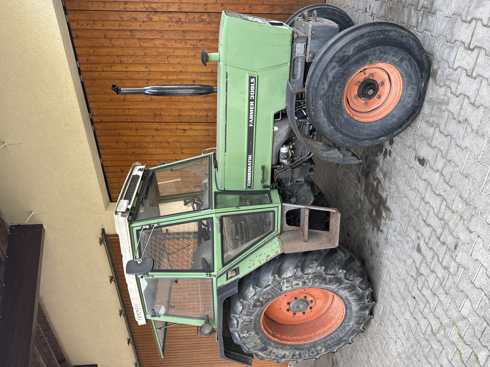 The image shows a green Fendt Farmer 309LS Turbomatik tractor parked on a paved surface. The tractor is positioned in front of a wooden garage door. The location is Riedlingen, Germany.