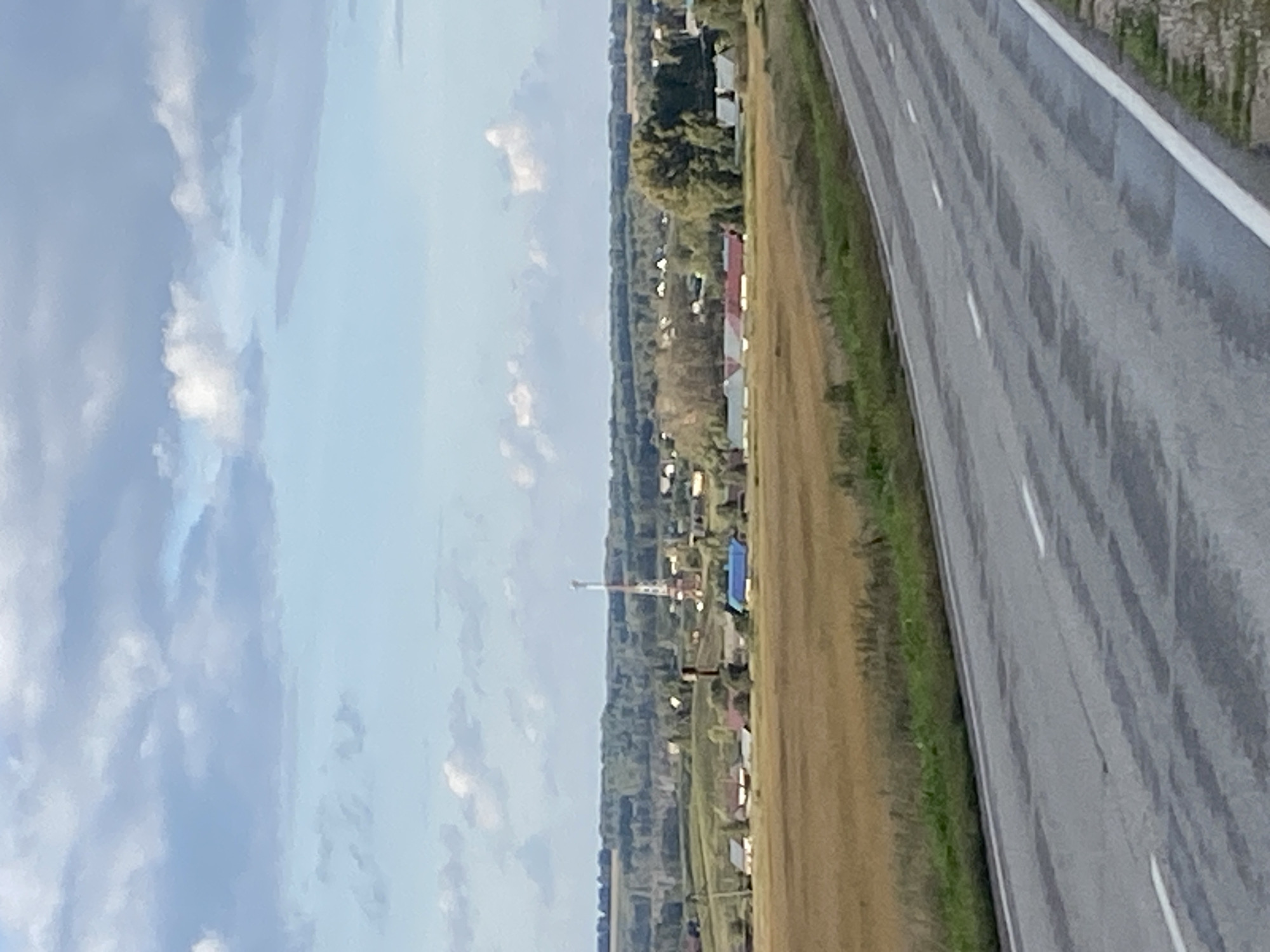 The image captures a rural landscape with a paved road in the foreground. The road has white lane markings and appears to be slightly wet. Beyond the road, a field of dry, golden grass stretches towards a village. The village consists of several houses with red and blue roofs, nestled amongst green trees. A tall, slender radio tower stands prominently in the village. The background features rolling hills covered in dense green forest. The sky is partly cloudy with patches of blue. The scene appears to be set in Медведское, Russia, given the architectural style of the village and the natural environment. No people are visible in the image.
