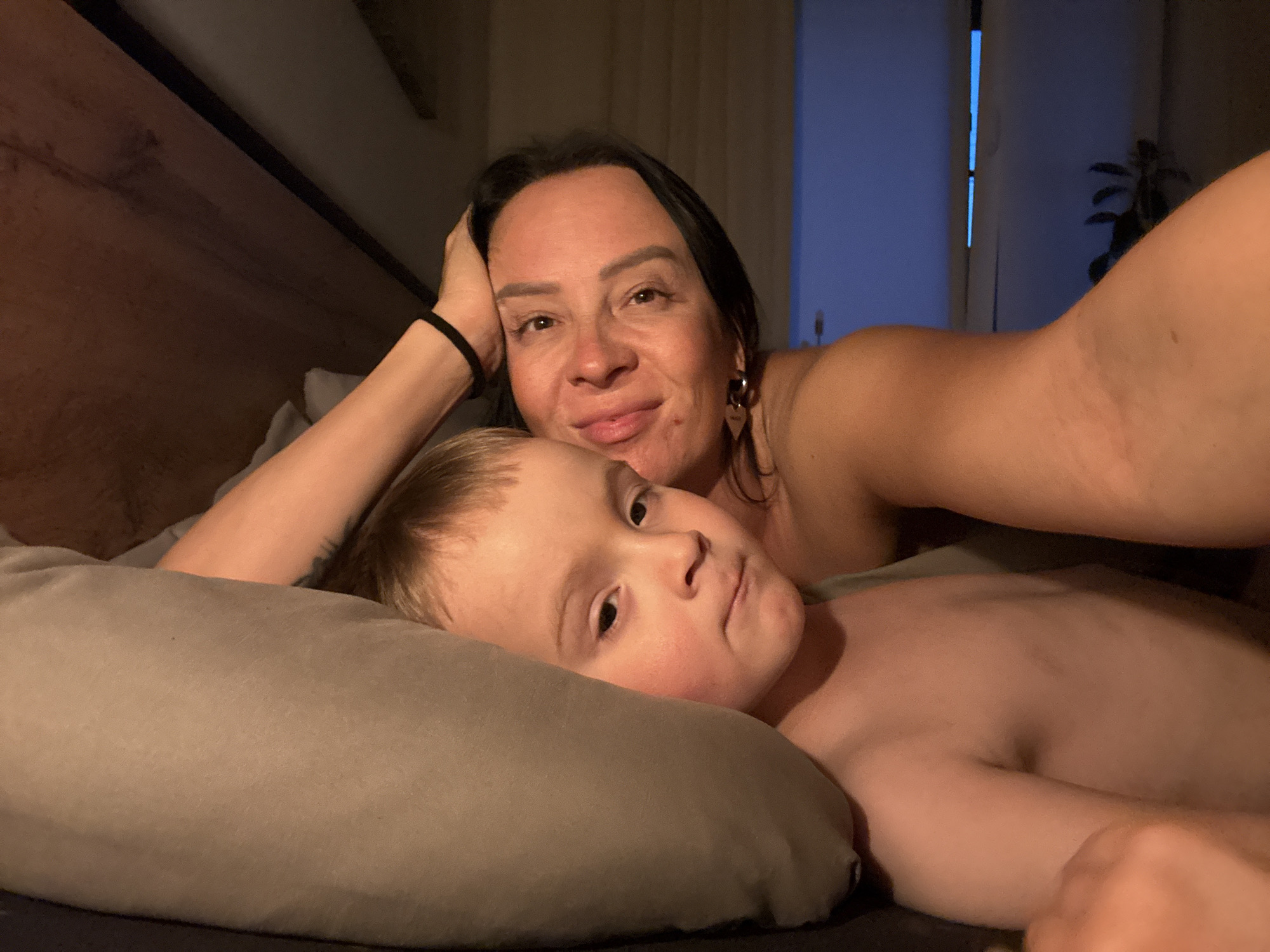 The image shows a woman and a young child lying in bed. The woman is fair-skinned with dark hair, and is resting her head on her hand. She appears to be taking a selfie. The child is lying on a pillow. They are indoors, possibly in a bedroom. The location is not discernible from the image. No information confirms this photo was taken in Chaikovsky, Russia.