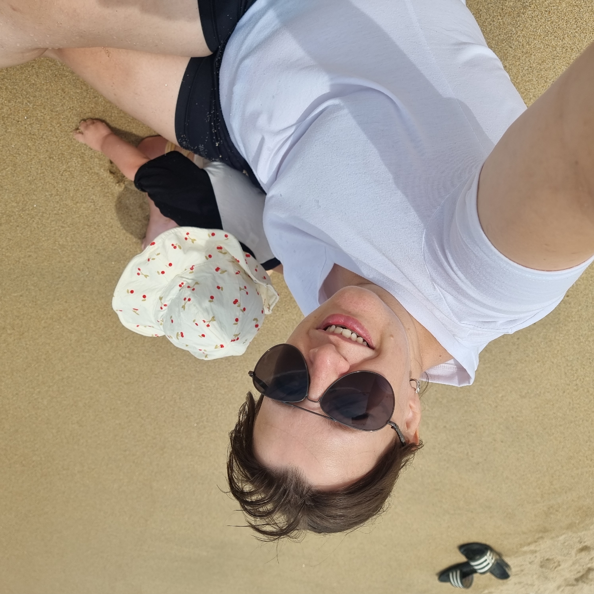 The image shows two people on a sandy beach. The person in the foreground is a woman wearing sunglasses, a white t-shirt, and black shorts. She is smiling and looking at the camera. To her right, a baby is sitting on the sand, wearing a white hat with a cherry pattern and a black and white outfit. In the background, a pair of black slides with white stripes are visible on the sand. The overall environment is a beach with fine sand and the faint presence of water at the edge of the frame, suggesting a coastal location. This scene is consistent with a typical recreational area in Gangneung-si, South Korea.