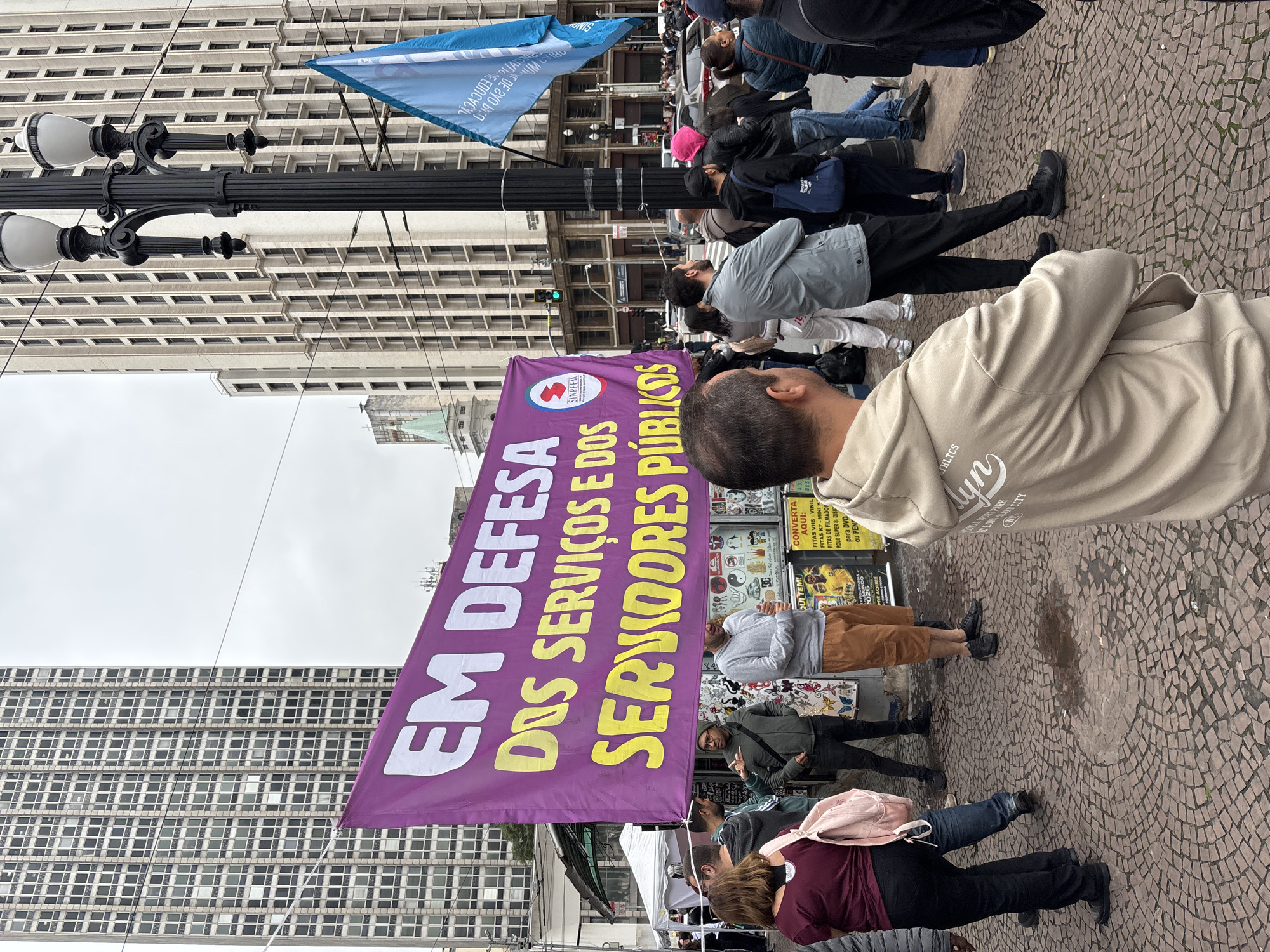 A demonstration takes place on a paved square in São Paulo, Brazil, in front of a tall, light-colored building with numerous windows. A purple banner with yellow lettering, partially visible, reads "EM DEFESA DOS SERVICOS E DOS SERVIDORES PUBLICOS." The banner also displays the name "SINPEEM." A light pole with ornate lamps stands beside the banner. People are visible, many standing, looking in various directions, while some carry additional banners. There are posters in the background. The sky appears overcast.
