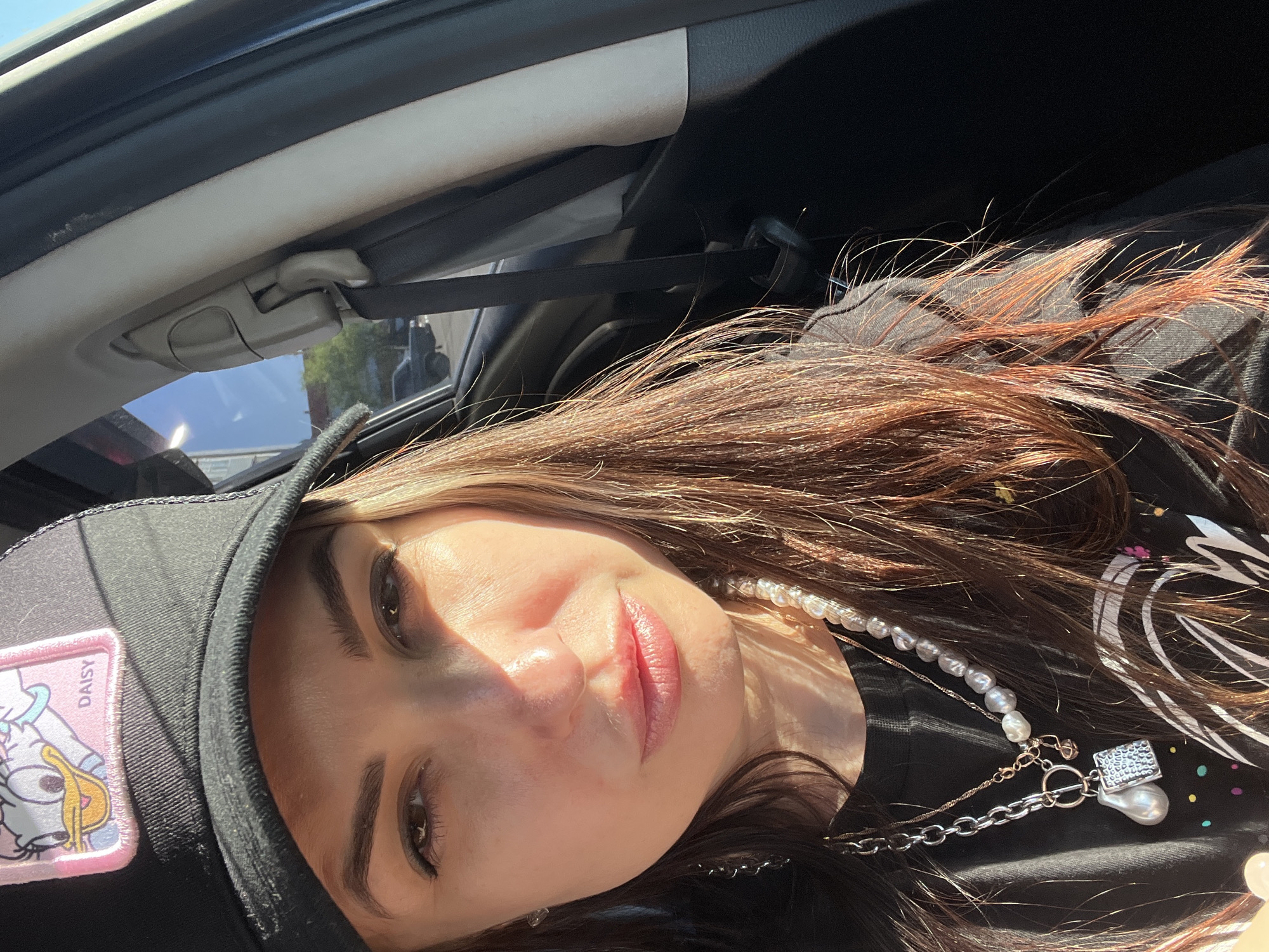 A young woman with shoulder-length brown hair with auburn highlights is in the passenger seat of a car. She is wearing a black baseball cap with a "Daisy Duck" patch and a pearl necklace with a pearl pendant. She is wearing a black shirt with a graphic design of white swirls and multicolored dots. The seatbelt is across her body. The location is Unknown, Unknown.