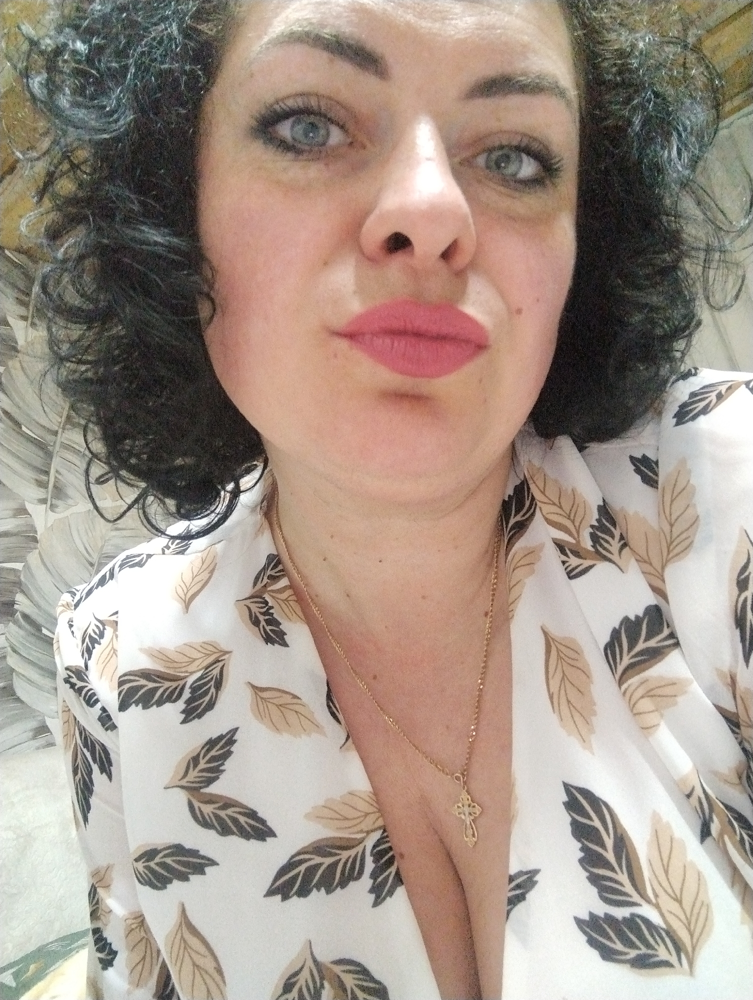 This is a close-up, head-and-shoulders portrait of a woman, captured indoors in Grishanki, Russia. She has dark, curly hair and prominent blue eyes, enhanced with eyeliner. Her eyebrows are dark and shaped, and her lips are painted with light pink lipstick, forming a slight pout. She looks directly at the camera with a neutral expression.
She is wearing a white top or blouse adorned with a pattern of light brown and dark brown/black leaves. Around her neck, she wears a delicate gold chain from which a small, ornate gold cross pendant hangs, resting on her chest.
The background appears to be an indoor wall, featuring a light-colored, possibly textured or patterned surface with abstract shapes resembling leaves or feathers in shades of grey and white. The lighting is soft and even, typical of an indoor setting. There is no visible text in the image.