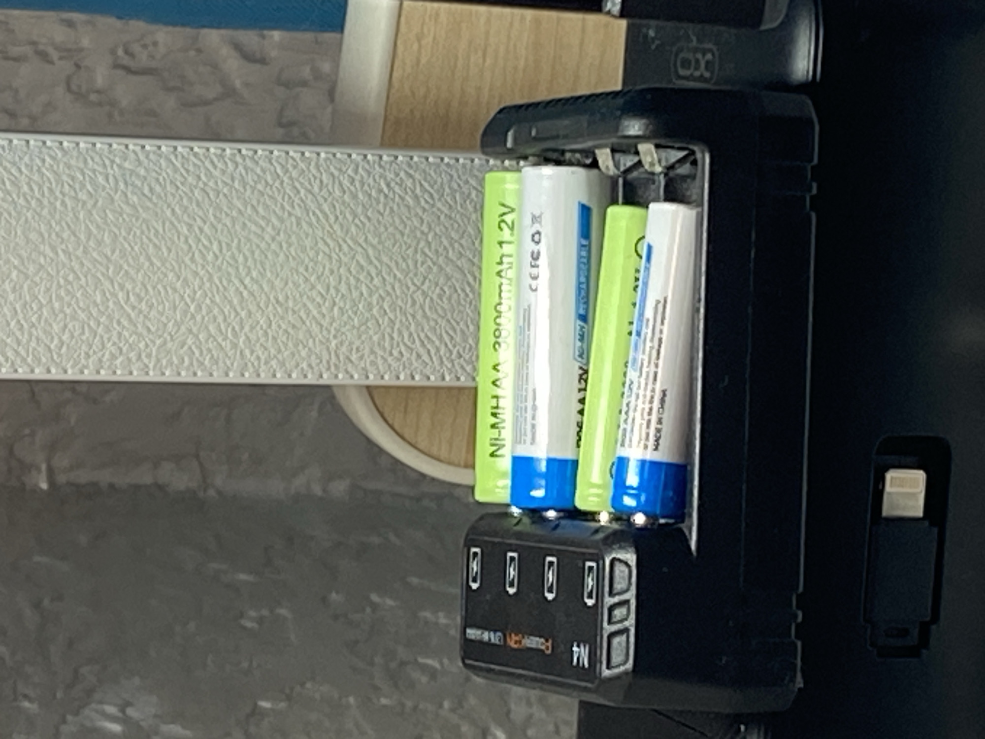 The image shows a black battery charger with four rechargeable AA batteries inserted. Two of the batteries are green and labeled "NI-MH AA 3800mAh 1.2V", and two are white and blue. The charger has four slots for batteries, each with an indicator light. In front of the charger, there is a small black object with a white connector, possibly a USB adapter. The charger and batteries are on a dark surface, and the background is out of focus, showing a textured grey surface and a portion of a white textured object. The lighting suggests an indoor environment. There is no indication of the location being Kryvyi Rih, Ukraine.