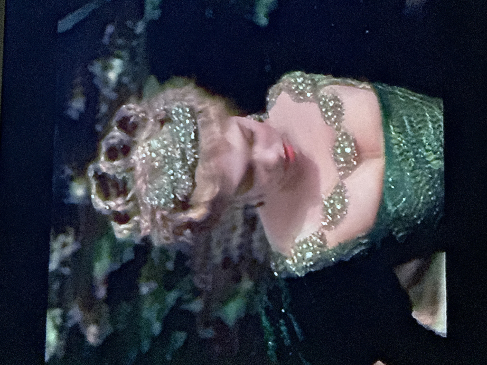 A person wearing a green dress with sparkling accents is lying down with their head tilted to the side. Their hair is styled with curls and adorned with similar sparkling decorations. The background is dark and out of focus, but appears to contain foliage. There is no information about the location in Mytishchi, Russia, so it is not included in the description.