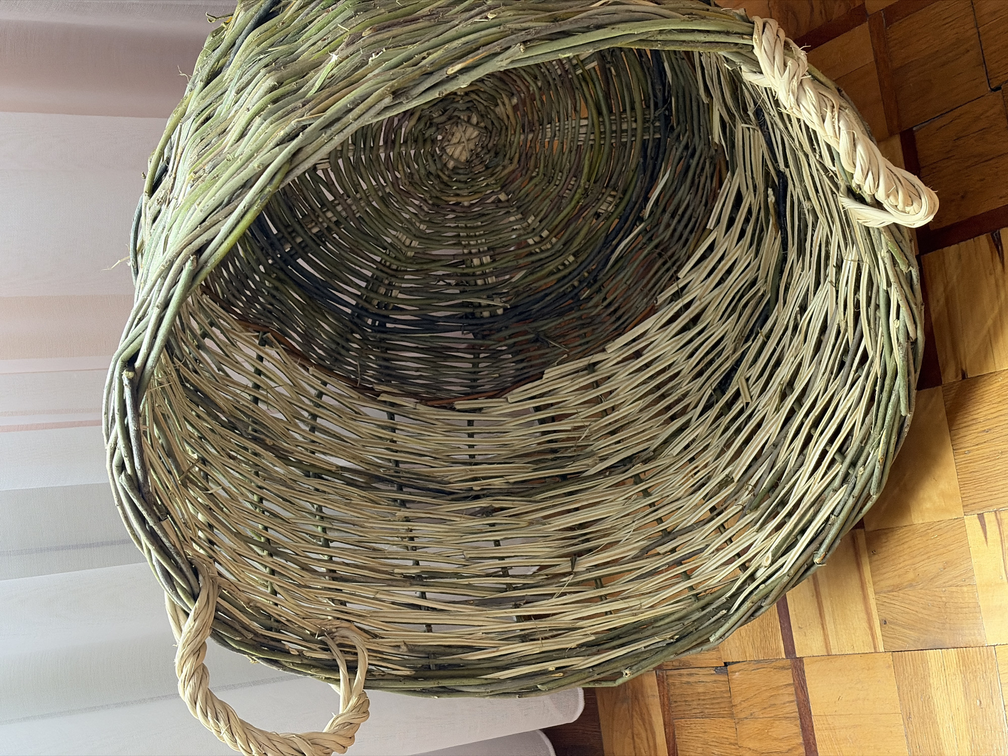 An overhead close-up shot shows a large, hand-woven basket lying on its side on a wooden floor. The basket is made of various natural materials, primarily thin, flexible branches and reeds, with some thicker, twisted rope-like handles. The outer layer of the basket consists of green branches of varying thickness, woven vertically and horizontally. Inside the basket, the weave changes to a spiral pattern made of lighter-colored reeds, with some darker sections. The basket is open and appears empty. The wooden floor beneath it has a parquet pattern, with alternating light and dark wood squares. A sheer, light-colored curtain is visible in the background, partially obscuring the top of the basket. The lighting is soft, casting gentle shadows. There are no people or animals in the frame. The scene appears to be indoors, likely in a domestic setting.