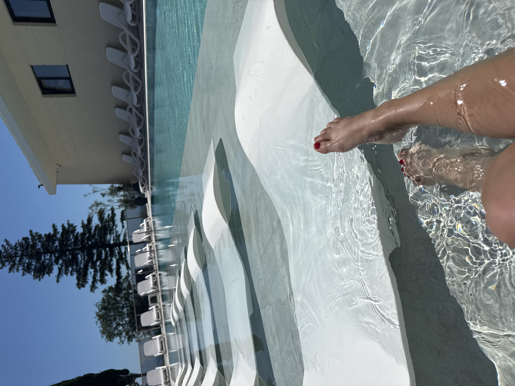 A person's legs and feet are submerged in shallow, clear water, with the person resting on a white, wave-shaped lounge chair. The lounge chair is positioned in an infinity pool, with more lounge chairs visible in the background. The pool is surrounded by a tiled area, and a building with windows and a beige facade is visible behind the pool. The sky is clear and blue, and a tall evergreen tree stands in the distance. The scene appears to be a resort or hotel setting.